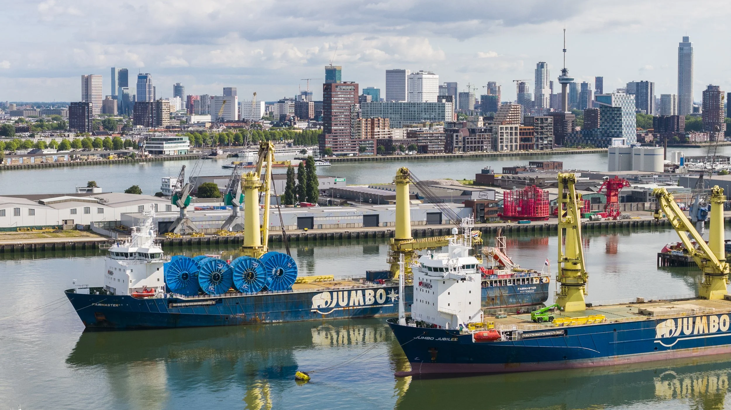 View of a harbor with ships and cranes, city skyline in the background, including tall buildings and a television tower.