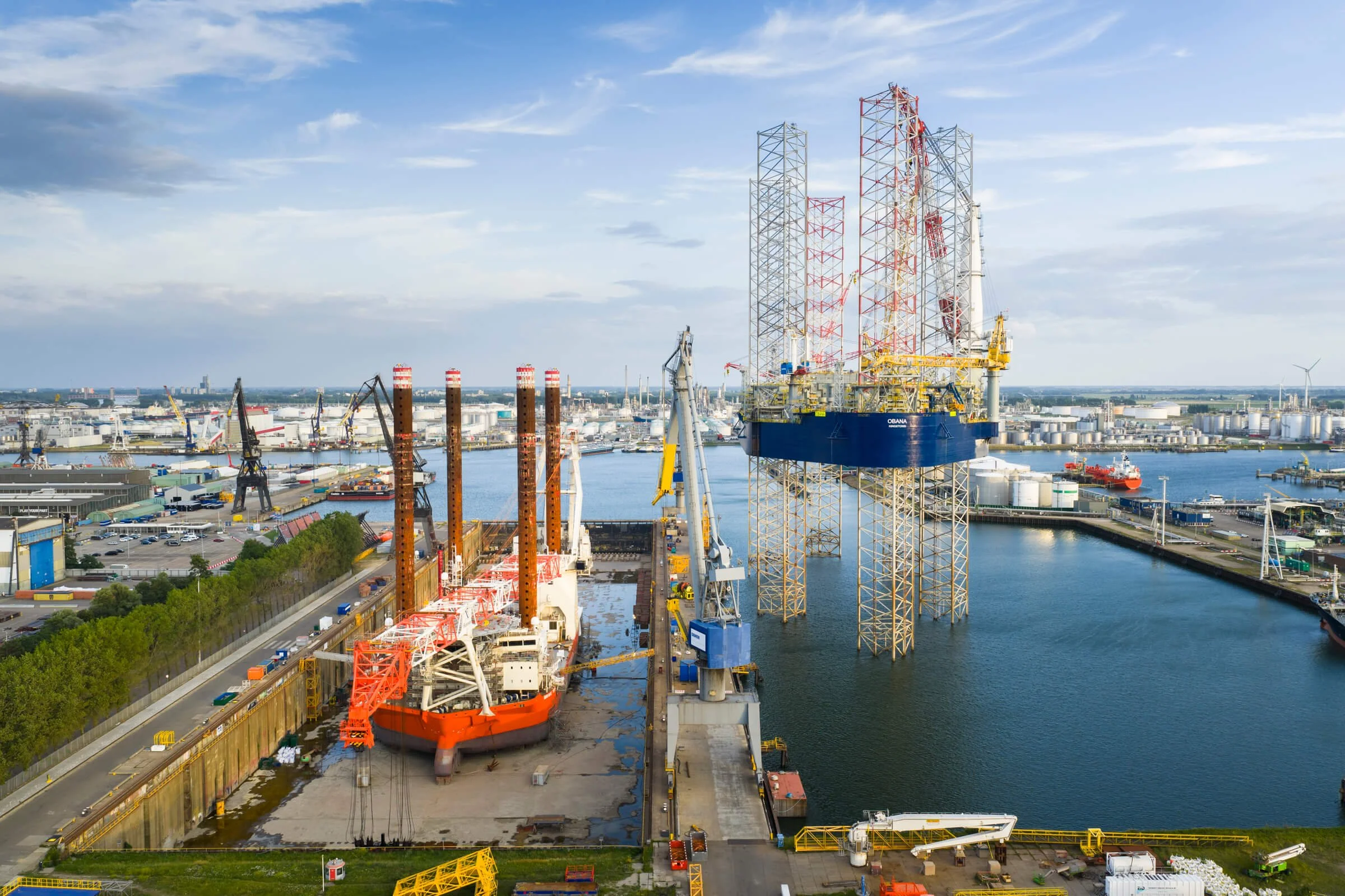 Industrial port scene with large drilling platform on water, ships, cranes, and storage tanks in the background.