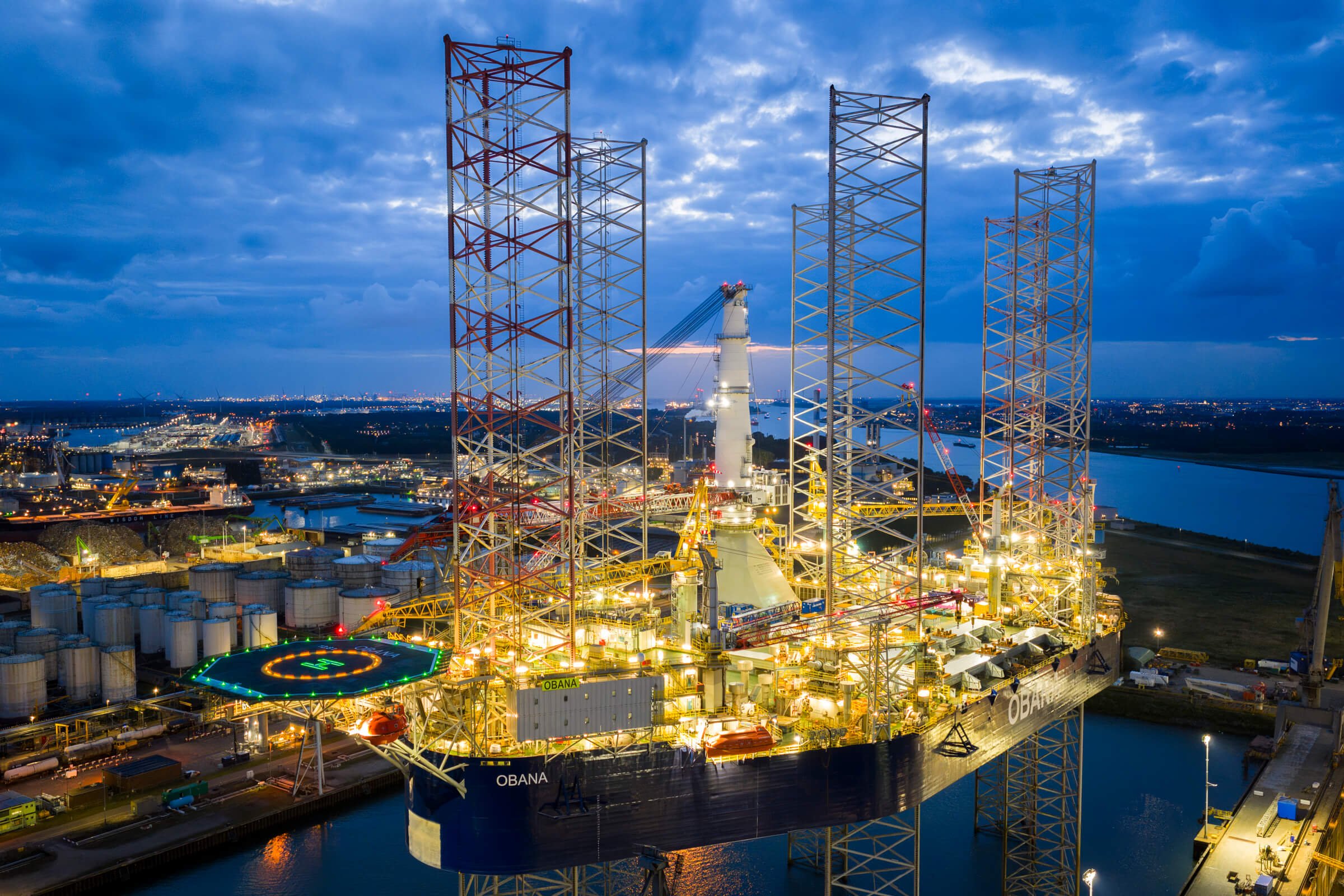 Oil drilling rig illuminated at night with a cityscape and river in the background.