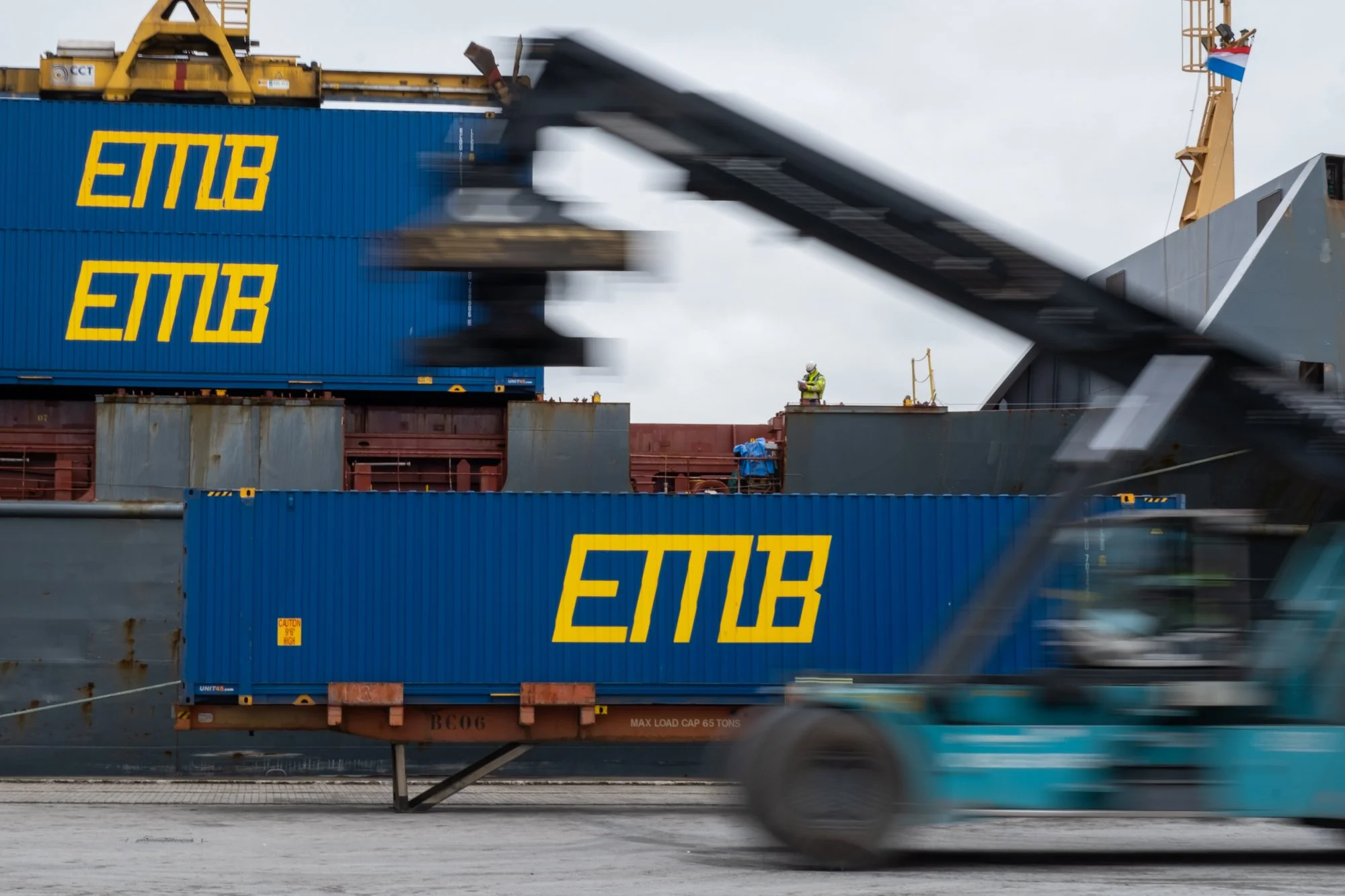 Cargo ship at port with blue and yellow containers labeled 'ETMB'. A forklift passes in front of the ship, and a worker in a safety vest and helmet stands on the deck.