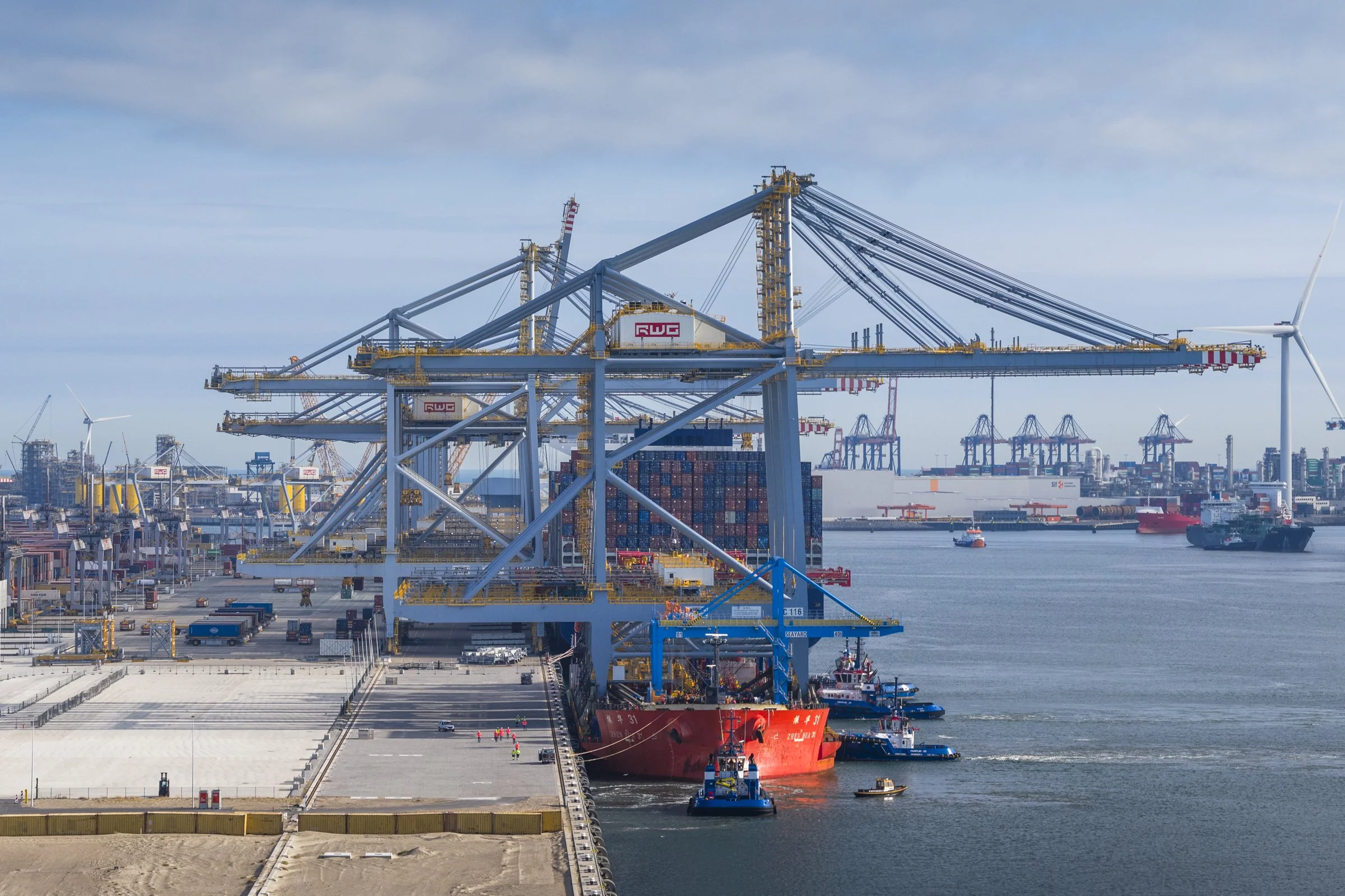 Port with large shipping cranes, containers, and ships docked on the water, with a wind turbine in the background.