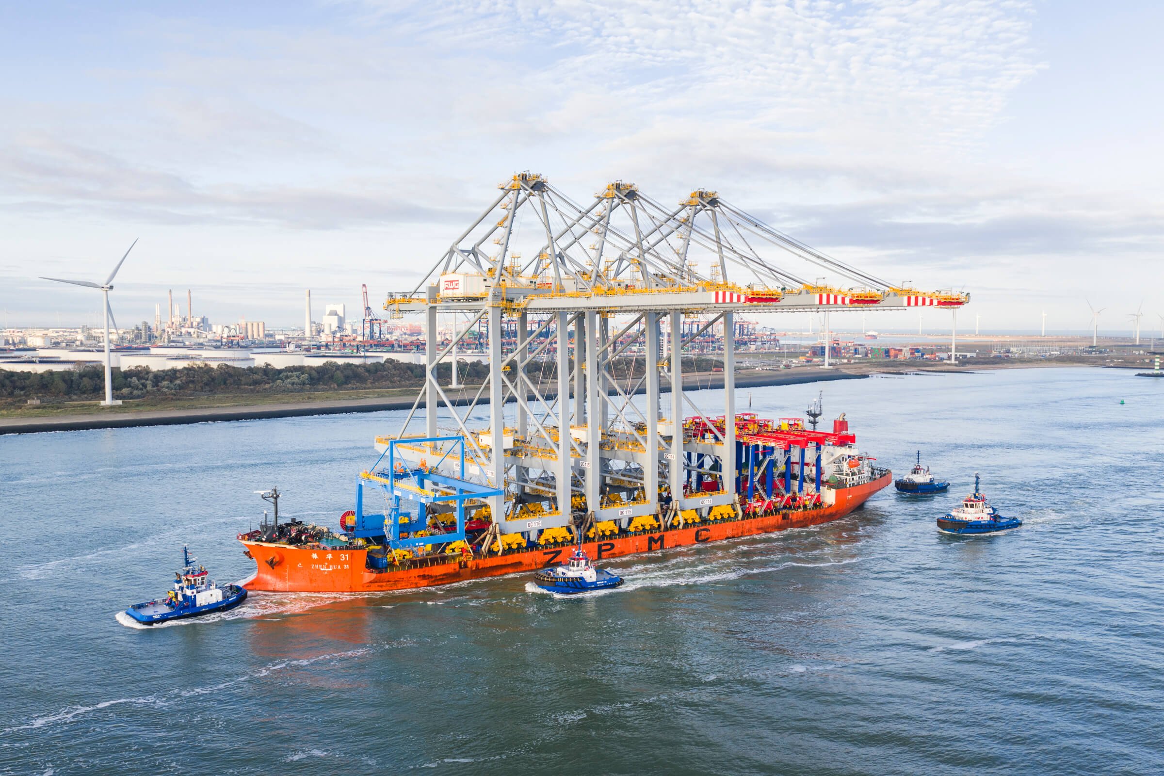 Cargo ship in a harbor being guided by three tugboats, with cranes and industrial facilities in the background.