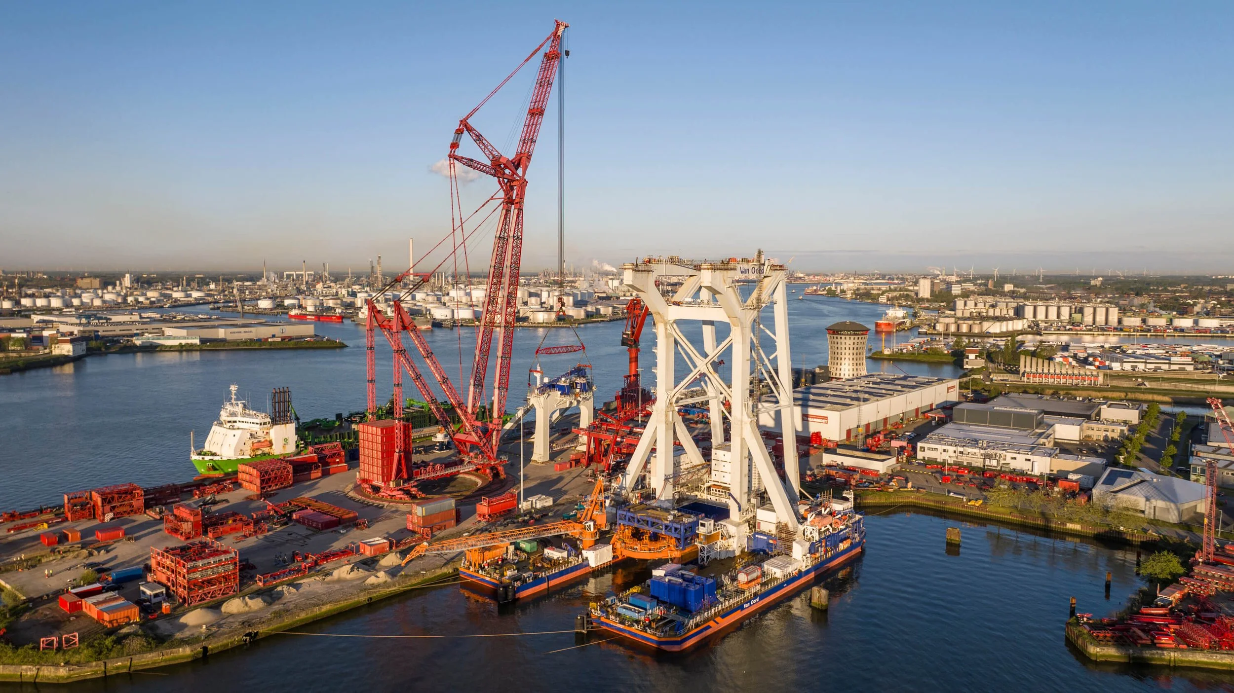 An aerial view of a busy port with large cranes, shipping containers, and ships on a body of water, with an industrial cityscape in the background.