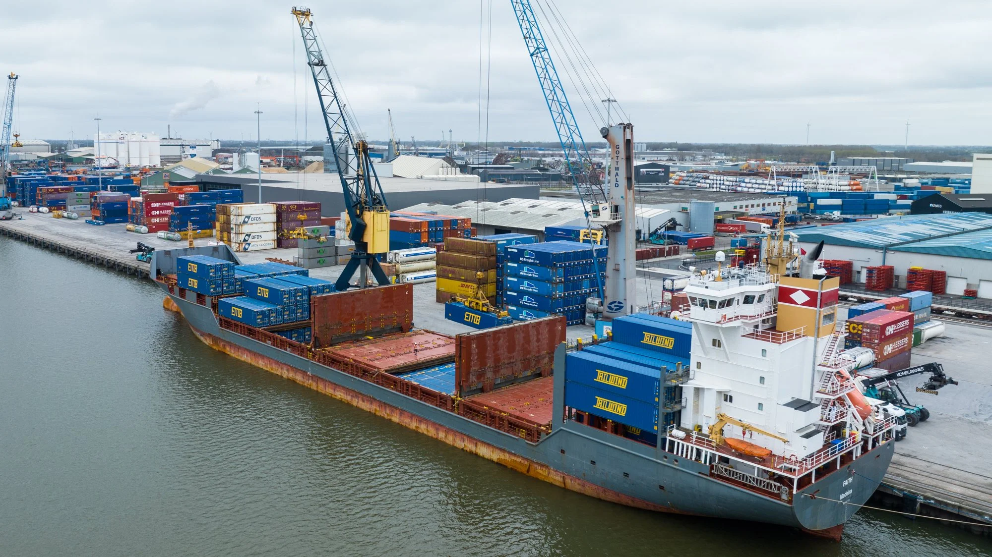 Cargo ship docked at a busy port with containers being loaded or unloaded using cranes, industrial warehouses in the background.