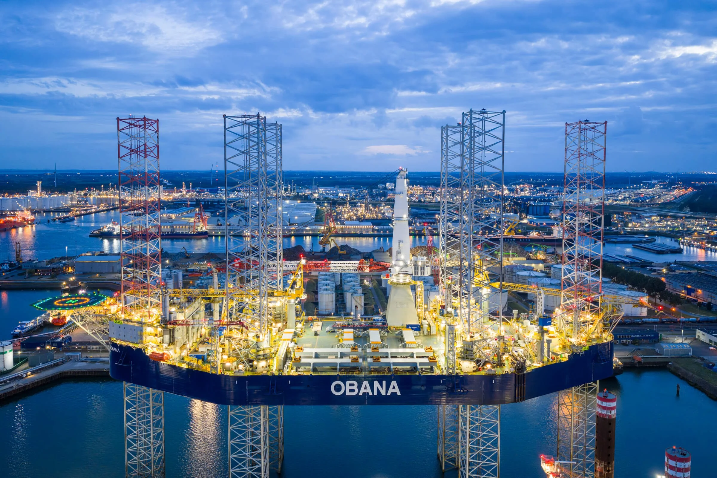 An offshore oil rig named OBANA illuminated at dusk, with multiple cranes and a cityscape in the background.