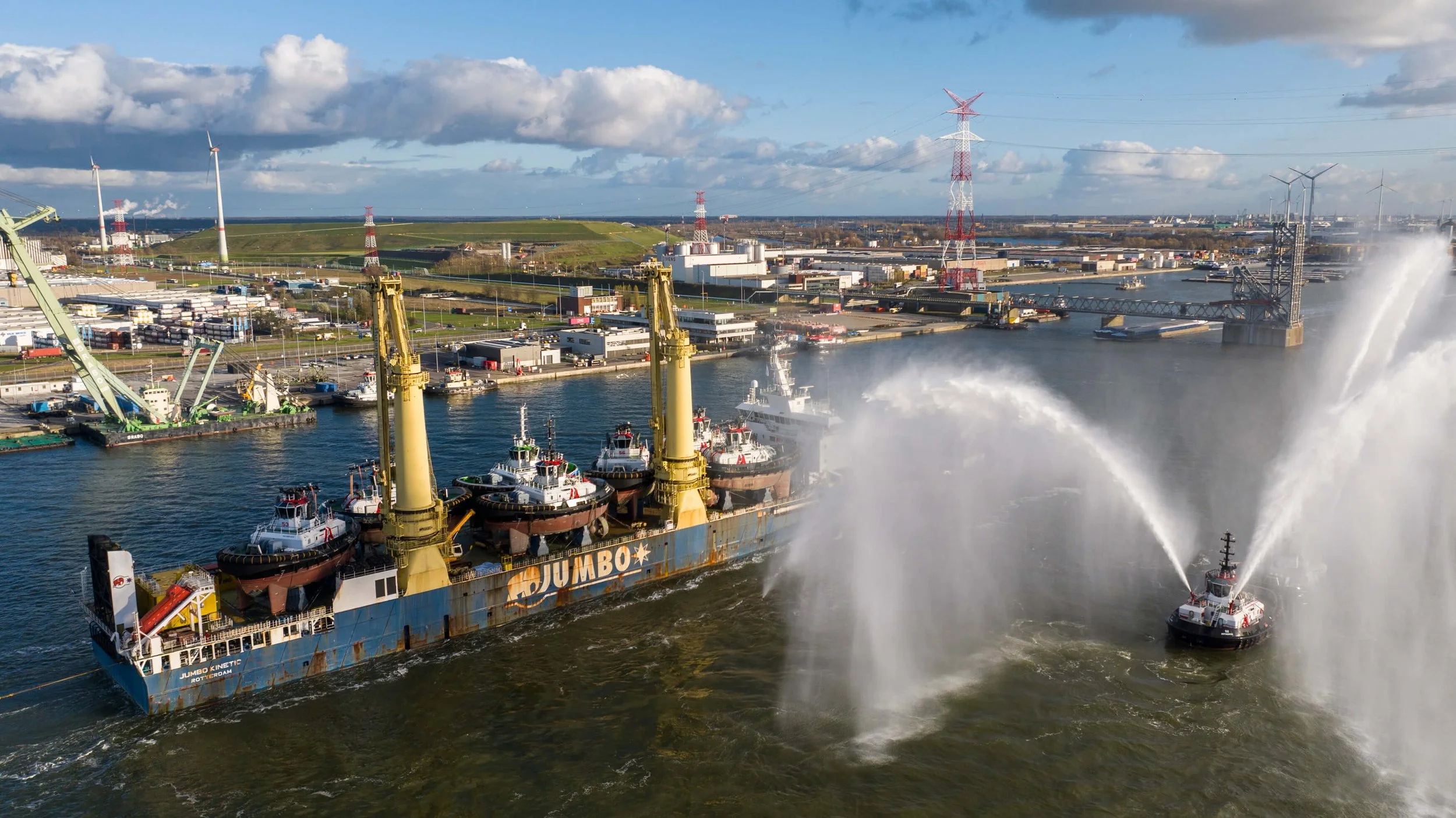 A large industrial vessel with yellow cranes on deck, carrying multiple tugboats, sailing in a harbor with water spray from a smaller vessel, with a background of wind turbines and industrial buildings.