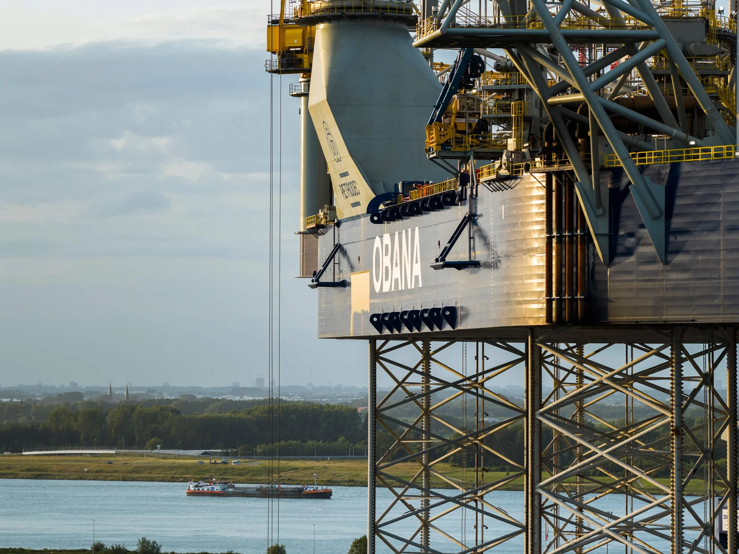 Offshore oil drilling platform named 'Obana' with a ship passing in the water below, in a landscape with trees and buildings in the distance under a cloudy sky.
