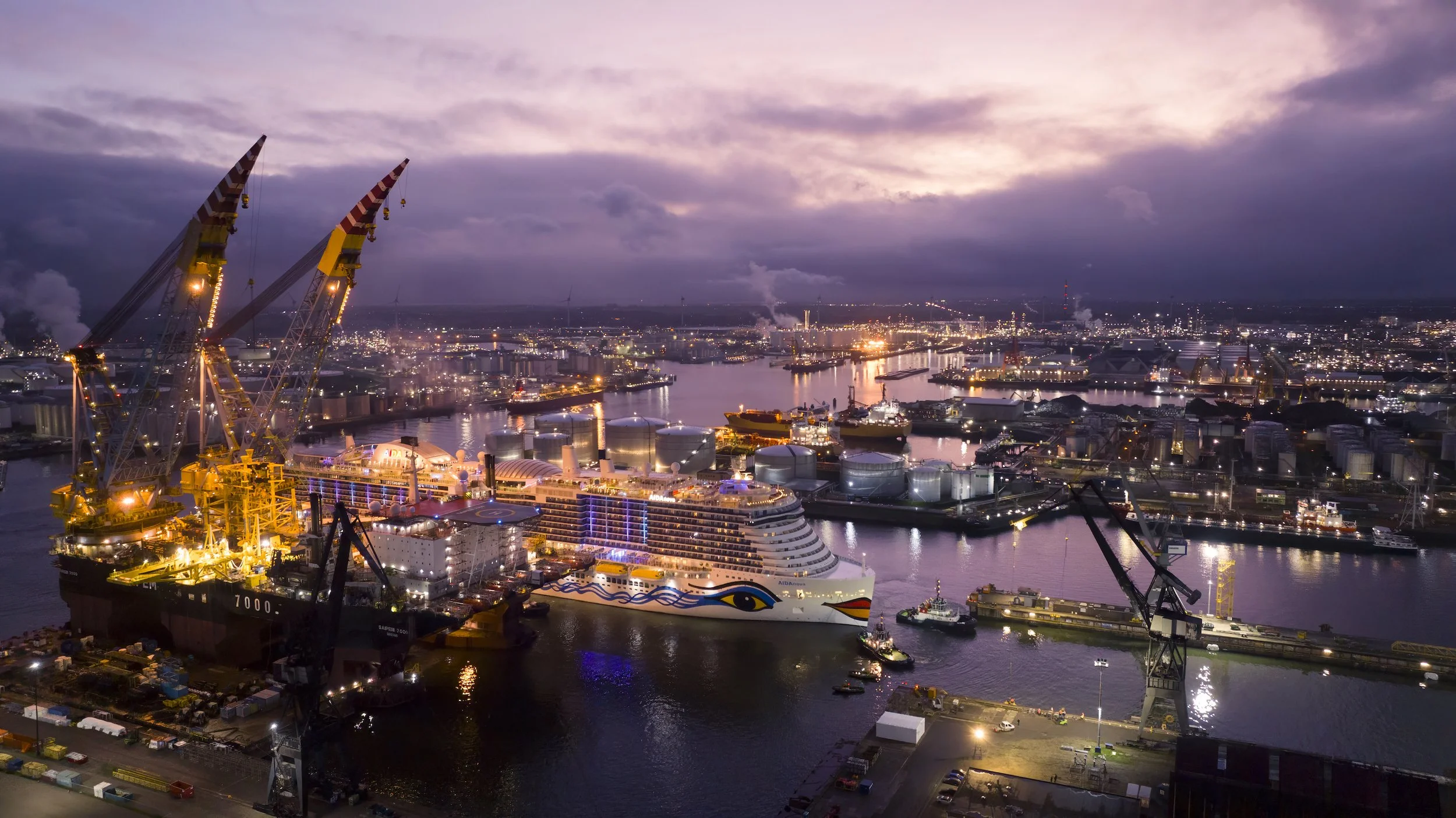 Nighttime view of a busy port with large cranes, a cruise ship, and industrial area illuminated by city lights and reflected on the water.