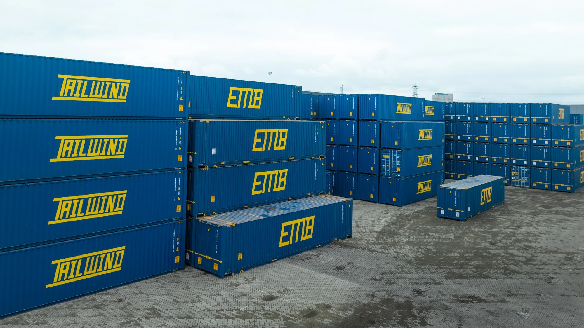 Multiple stacked blue shipping containers with yellow 'TALWIND' and 'ETMB' logos in an outdoor storage yard, with a cloudy sky and industrial background.