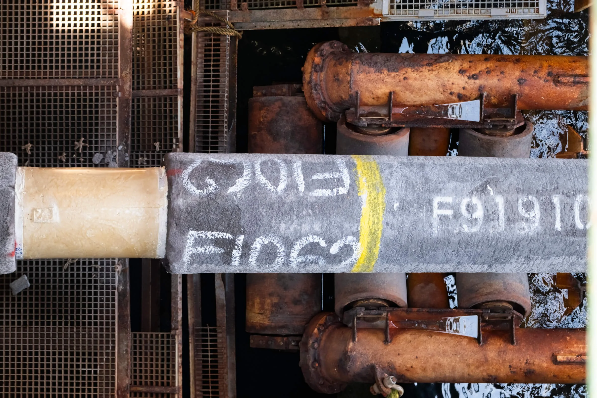 View from above showing a large concrete pipe with painted markings, rusted metal rollers, and a metal grate surface.