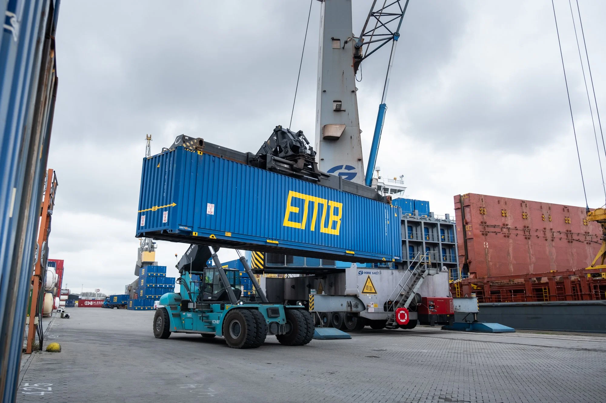 A container handling operation at a port with a large crane lifting a blue shipping container marked 'EMAS' and a forklift nearby.