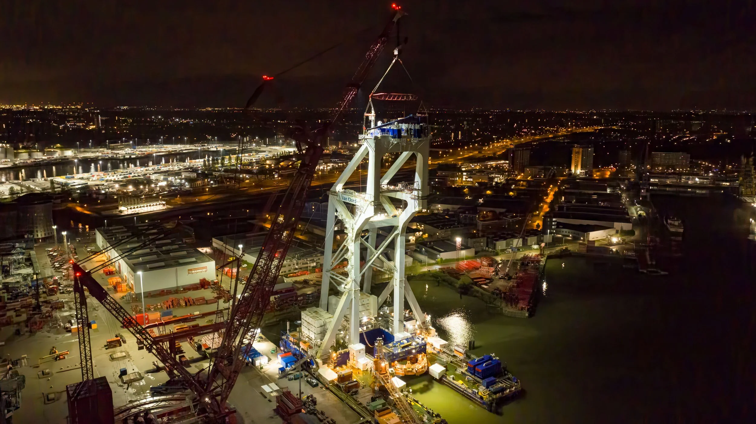 Nighttime view of a large bridge under construction with cranes and construction equipment, illuminated by floodlights, over a waterway in an urban area.