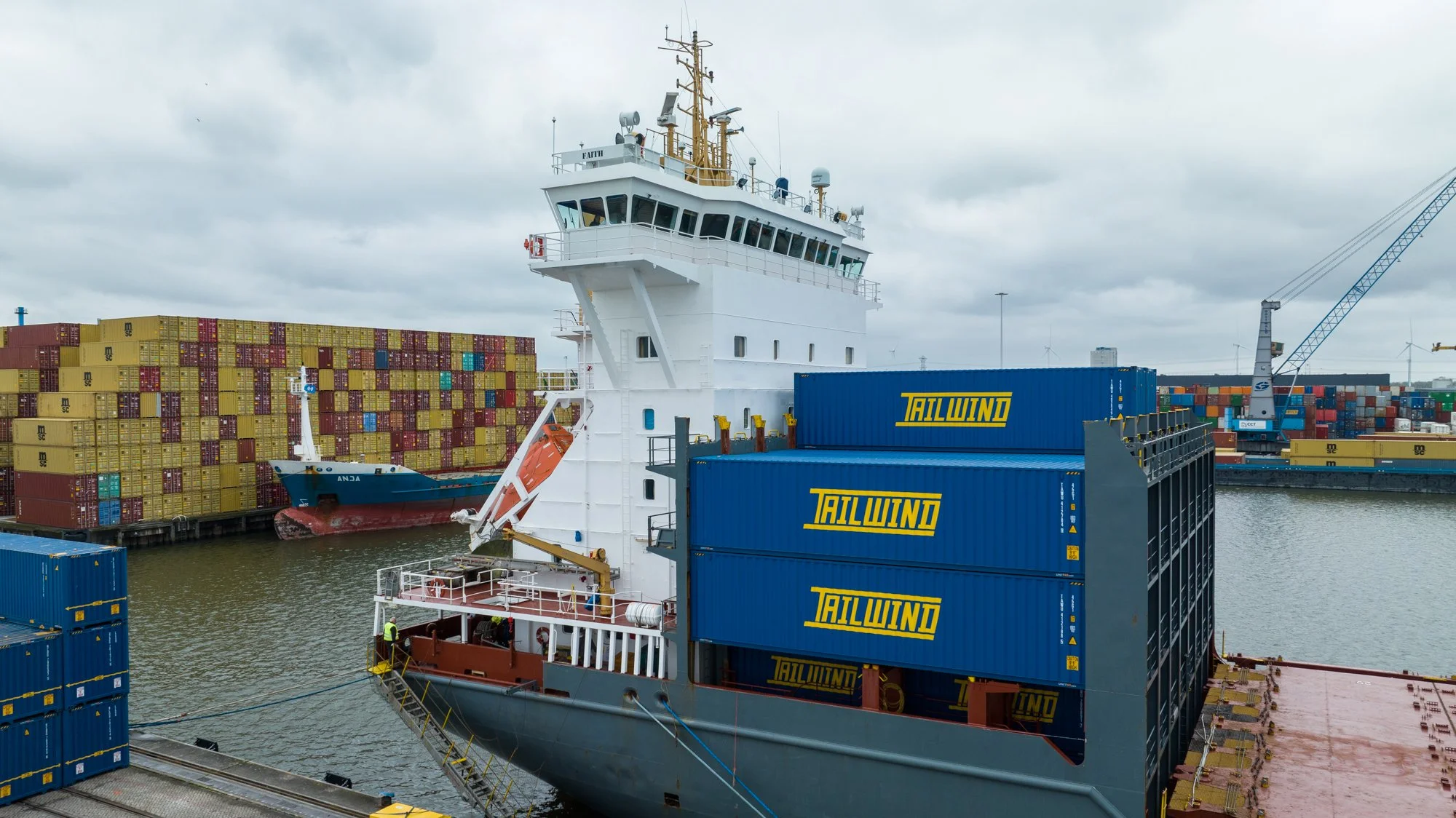 A cargo ship docked at a port with many stacked shipping containers, some labeled 'TALWIND'. The ship's bridge and control tower are visible, and there are workers on the deck. The background shows an industrial port with cranes and more containers.