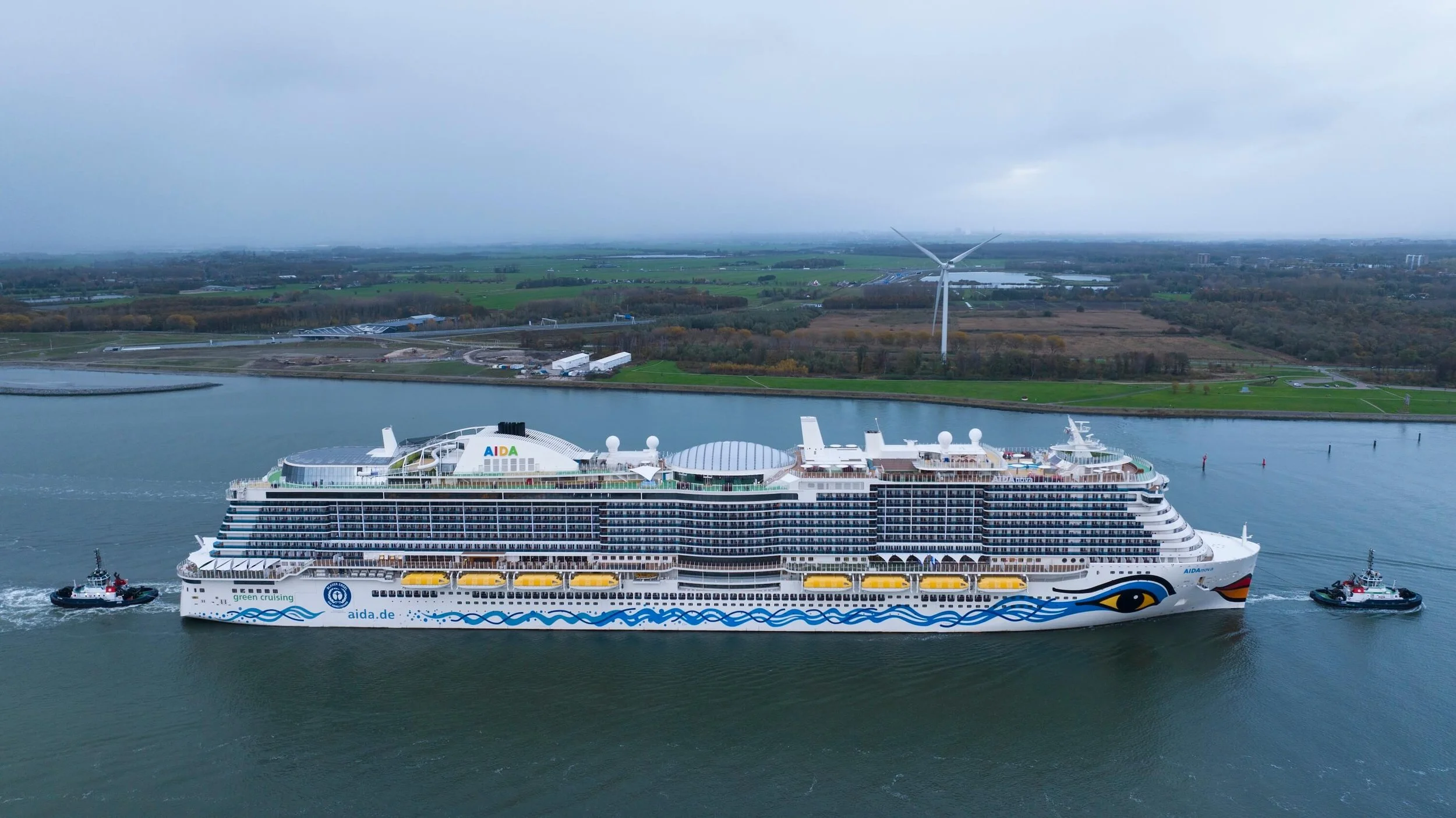 A large cruise ship with the word 'AIDA' on top, sailing on a river with tugboats on either side, surrounded by greenery and wind turbines in the background.