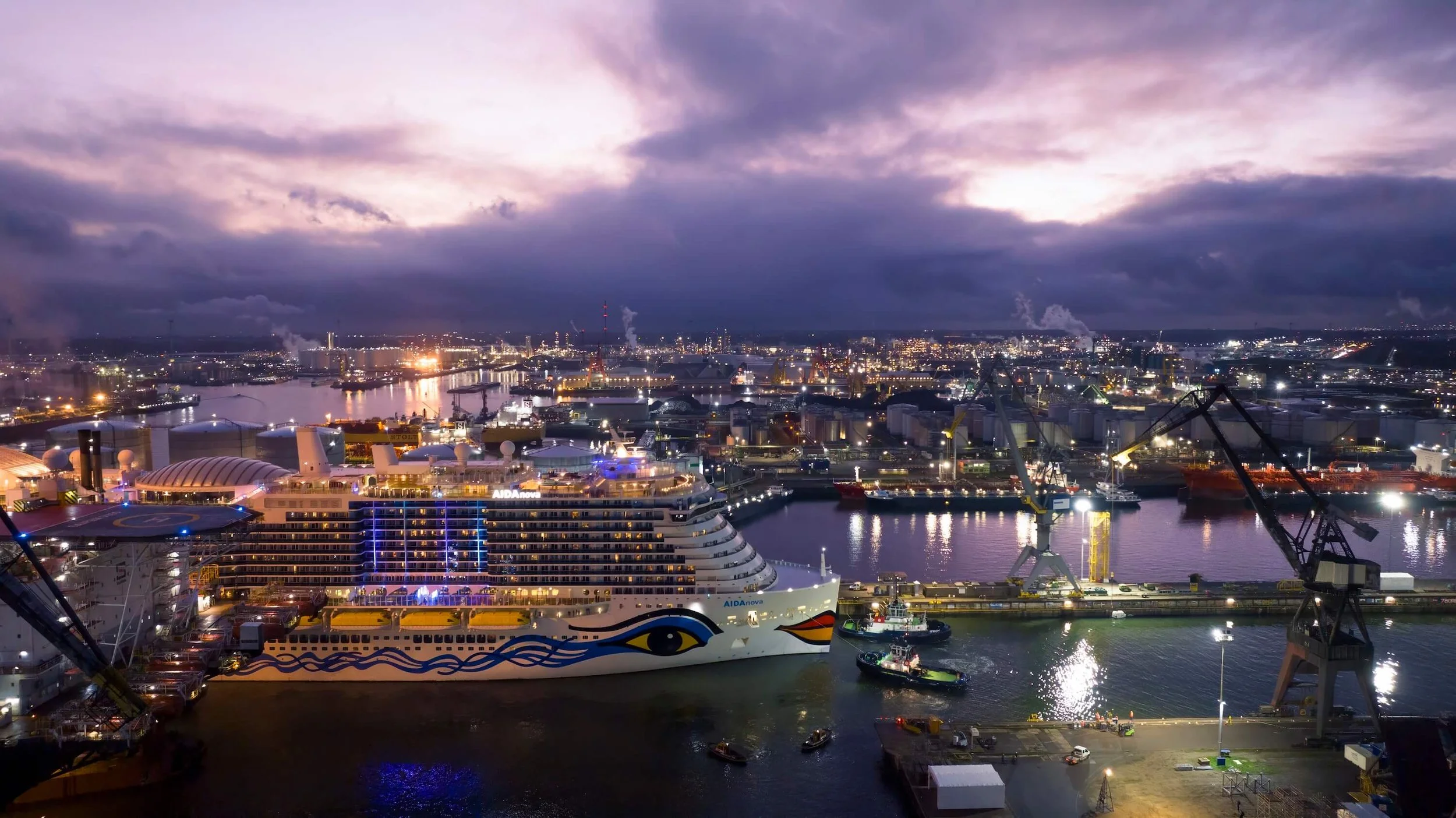 Night view of a city port with a large cruise ship decorated with colorful face-like artwork, cranes, and industrial buildings with city skyline and cloudy sky in the background.