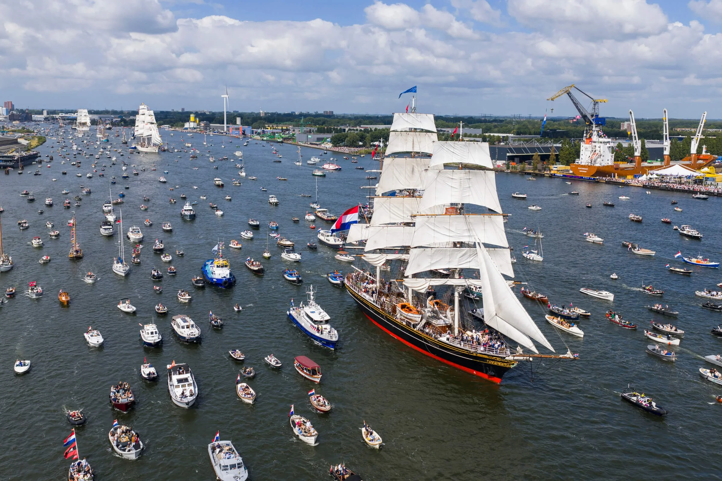 A large sailing ship with white sails navigating through a harbor filled with numerous small boats and yachts under partly cloudy skies.