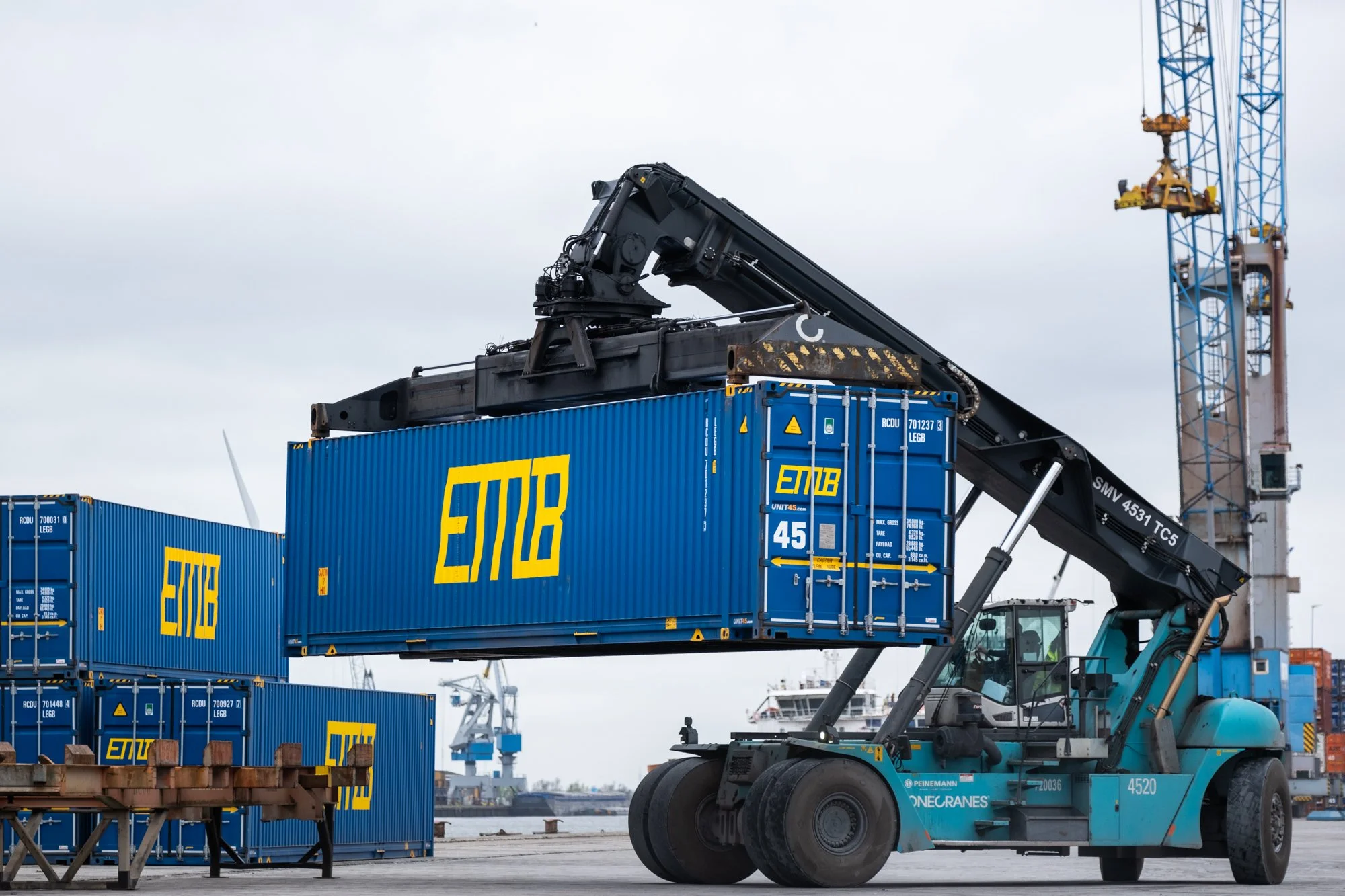 A forklift lifting a blue shipping container at a port with cranes in the background.