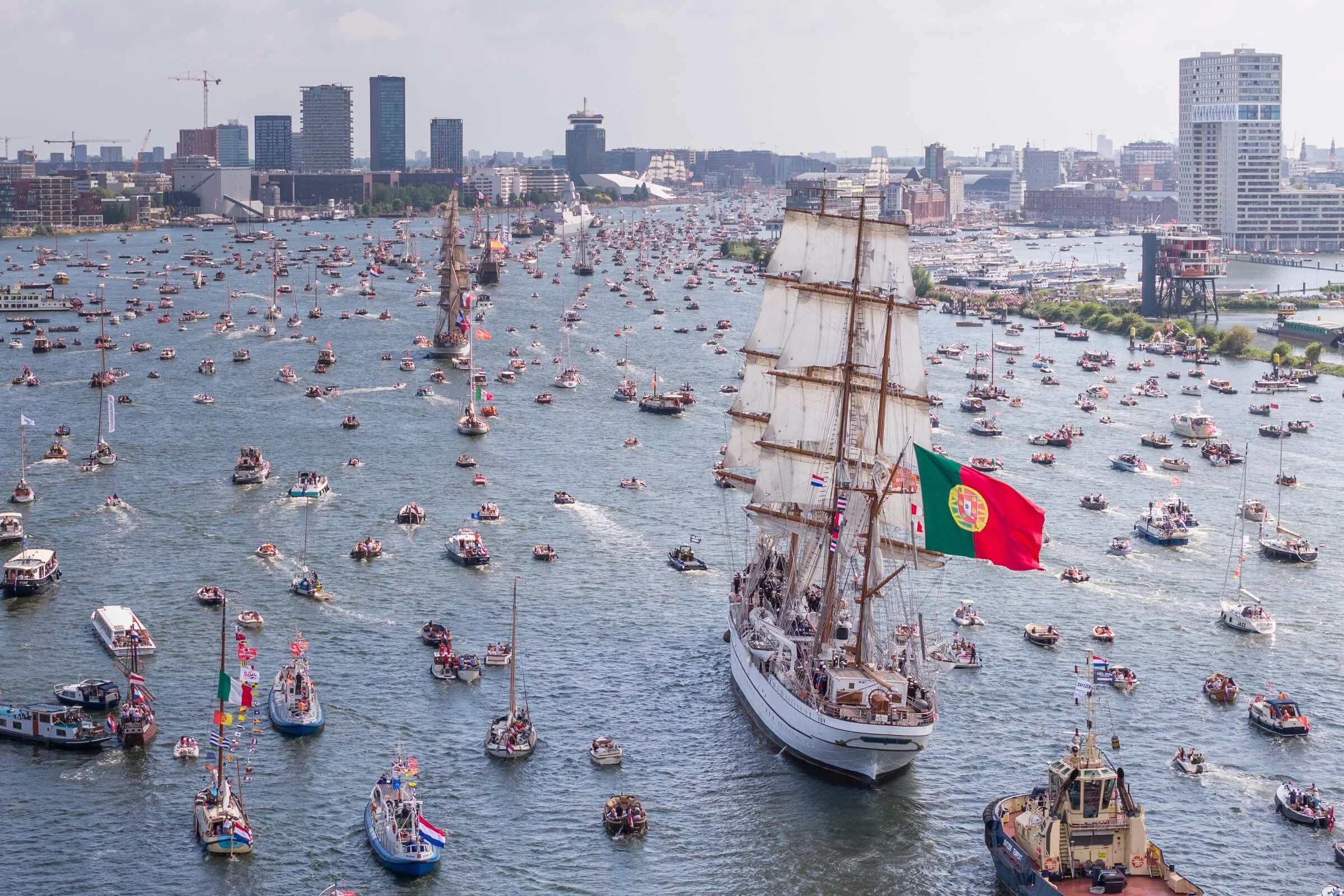 A large sailing ship with a Portuguese flag leading a fleet of smaller boats on a river in a city with tall buildings in the background.