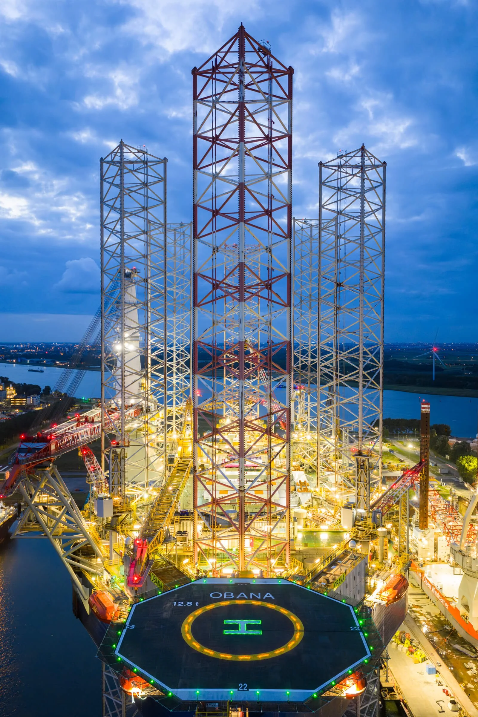 Nighttime view of an offshore oil platform with a helipad and towering red and white steel structures against a partly cloudy sky.
