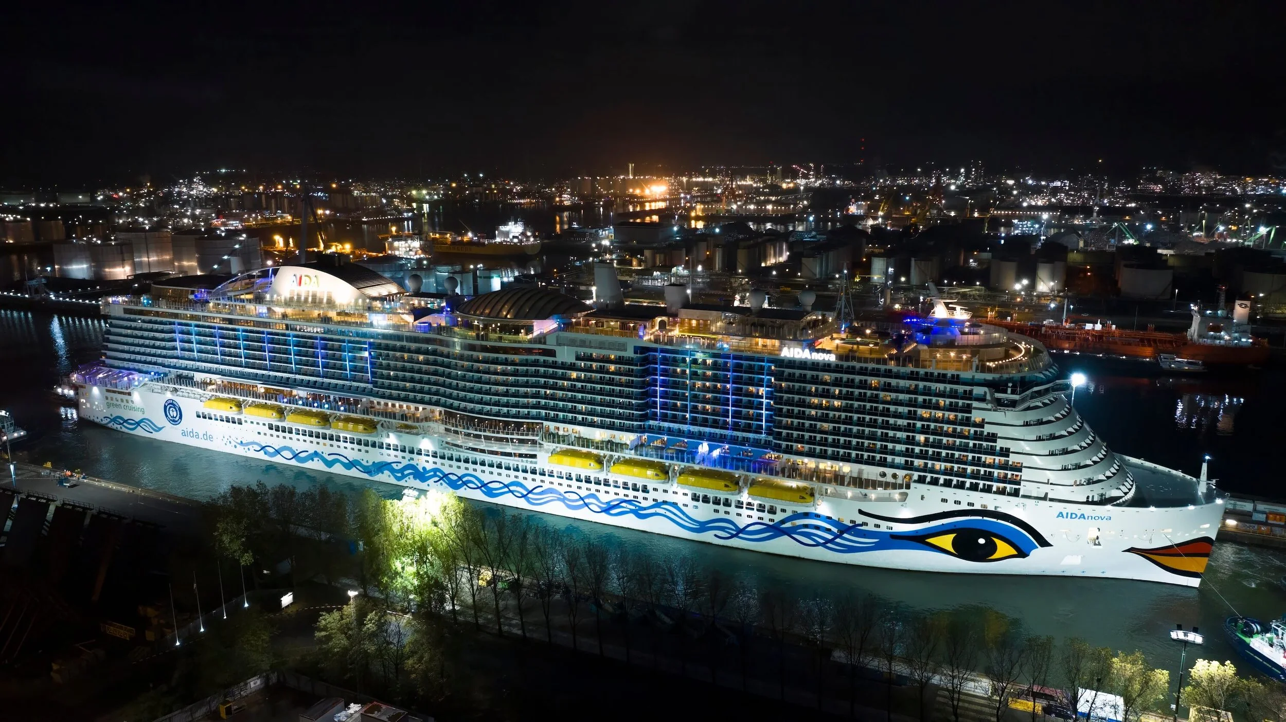 Nighttime aerial view of the illuminated cruise ship AIDAnova docked at the port, with city lights and industrial area in the background.