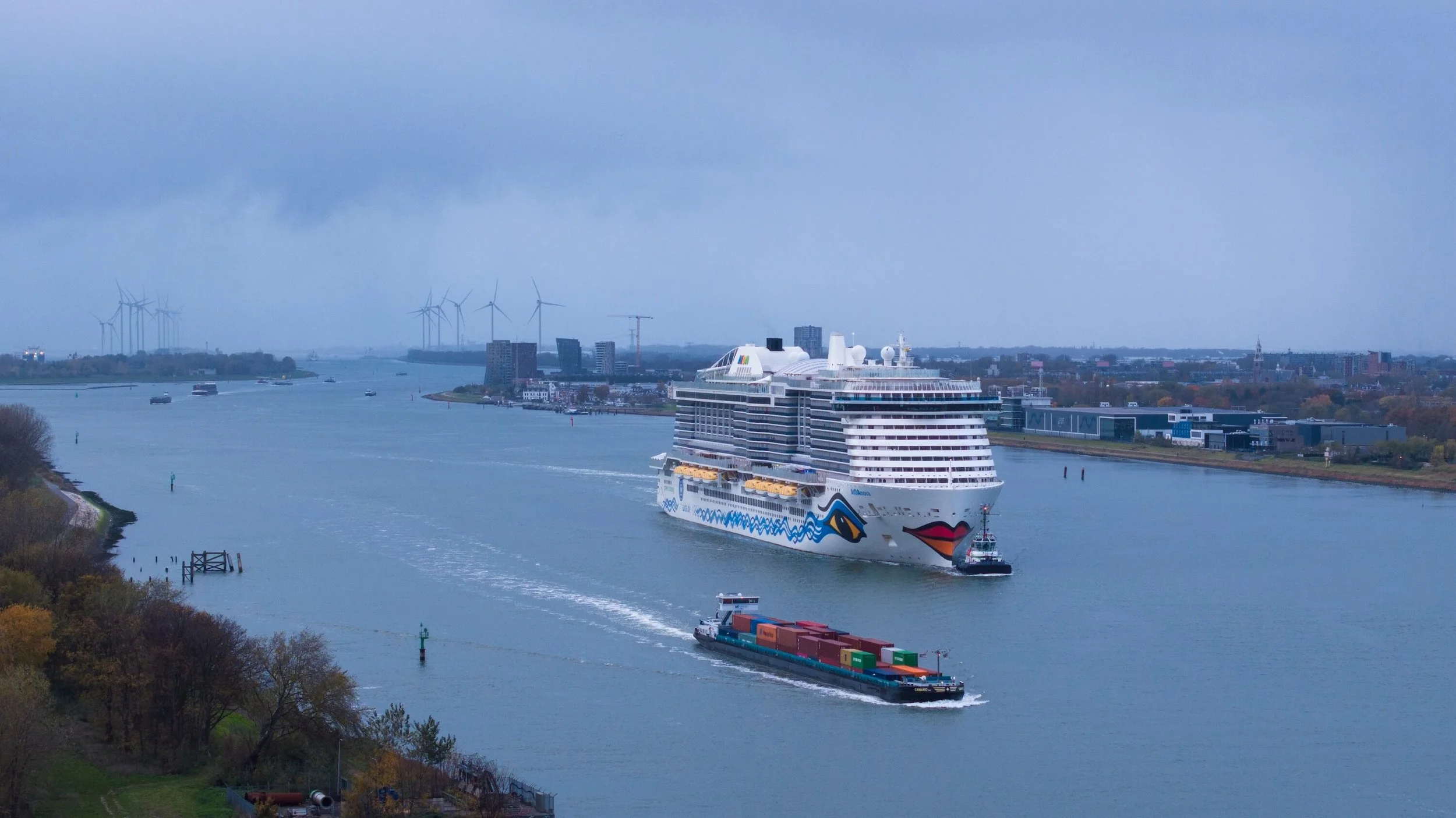 A large cruise ship with a face design, including red lips and painted eyes, sailing through a river with a cargo boat nearby. The city skyline and wind turbines are visible in the background on a cloudy day.