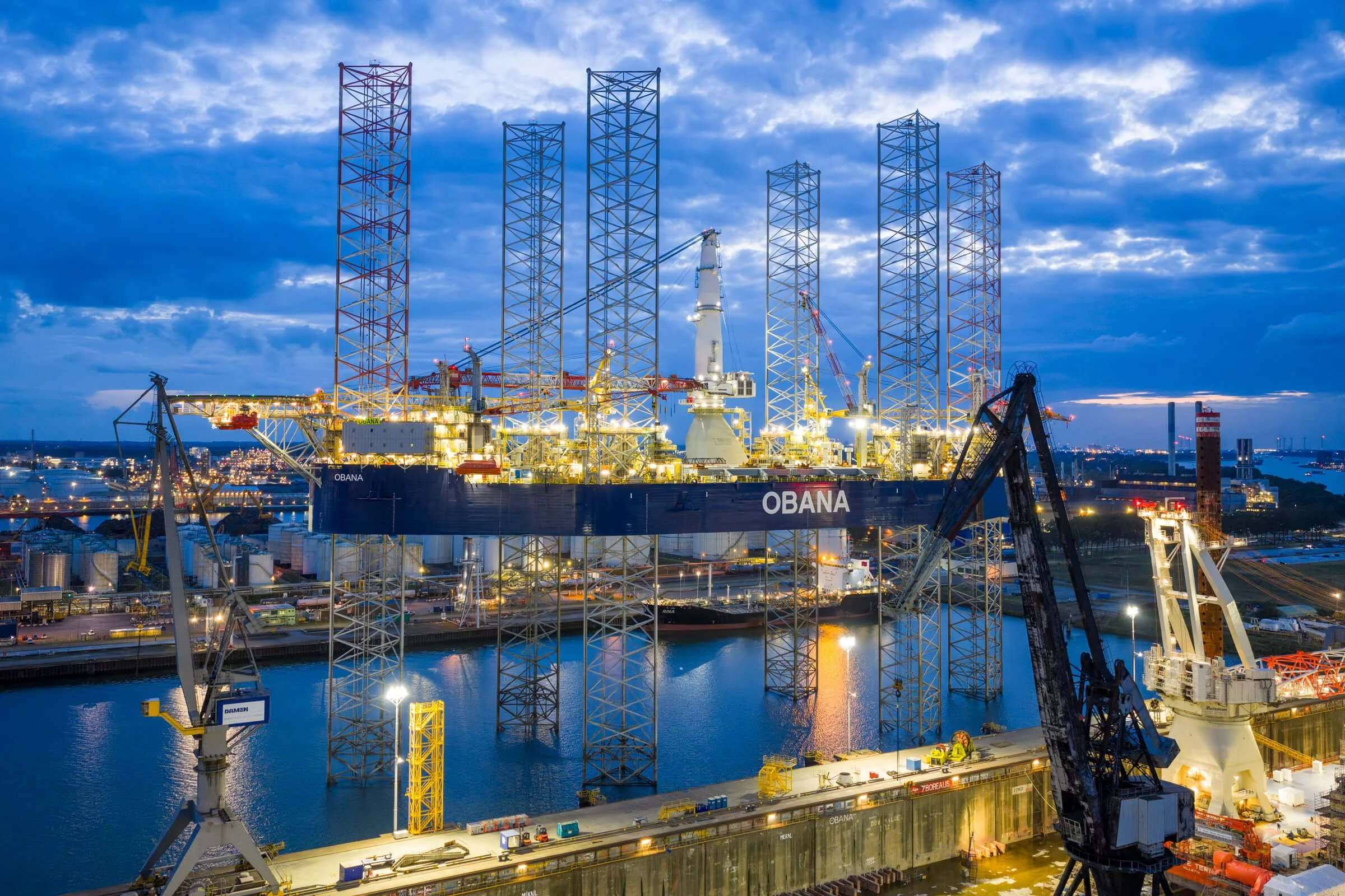 Offshore oil platform named OBANA with cranes and support vessels docked nearby, illuminated at dusk with a city and sky in the background.