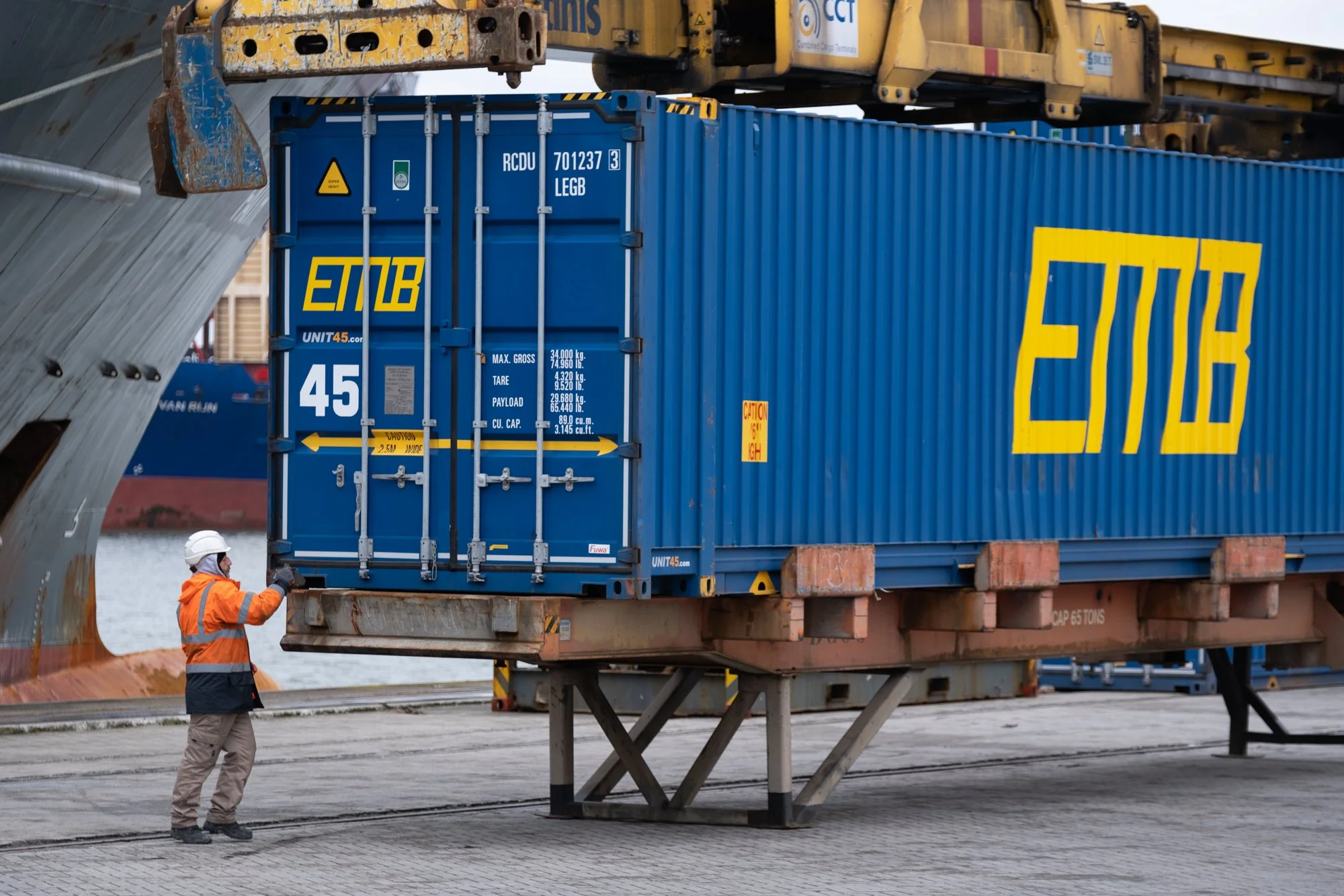 A worker in safety gear, including a helmet and orange safety vest, standing next to a forklift lifting a blue shipping container with yellow marking at a port.