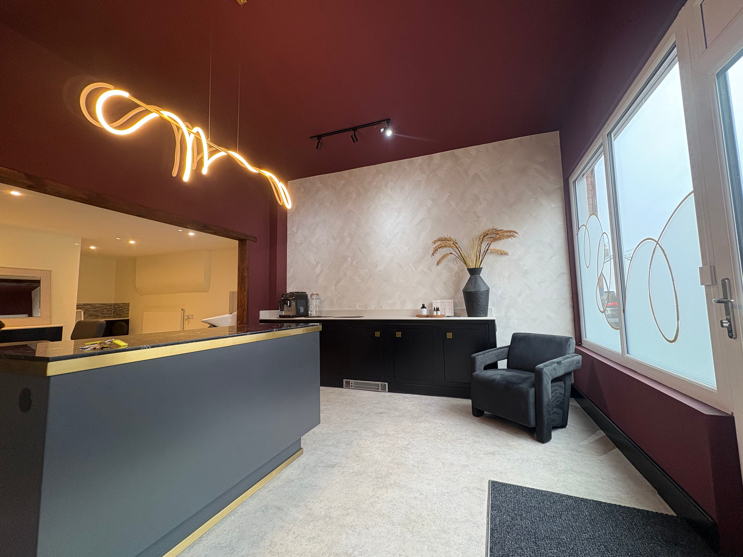 Modern interior lounge area with a black armchair, large window with circular decals, a black sideboard with a large vase and dried grass, maroon and beige walls, a unique yellow light fixture, and a partial view of a kitchen with a bar counter.