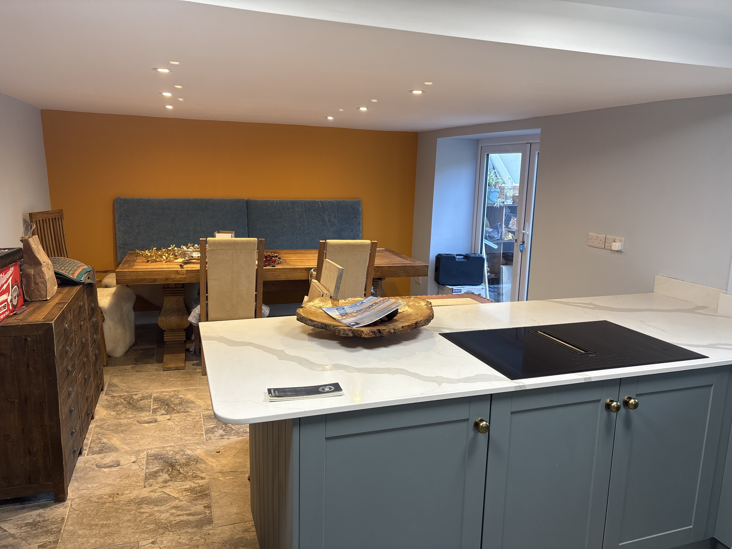 Modern kitchen with white marble countertop, gray cabinets with gold knobs, and a wood dining table with beige chairs. There is a yellow accent wall, and a sliding glass door leading outside. Decorative bowl on the counter and various items on the table and sideboard.