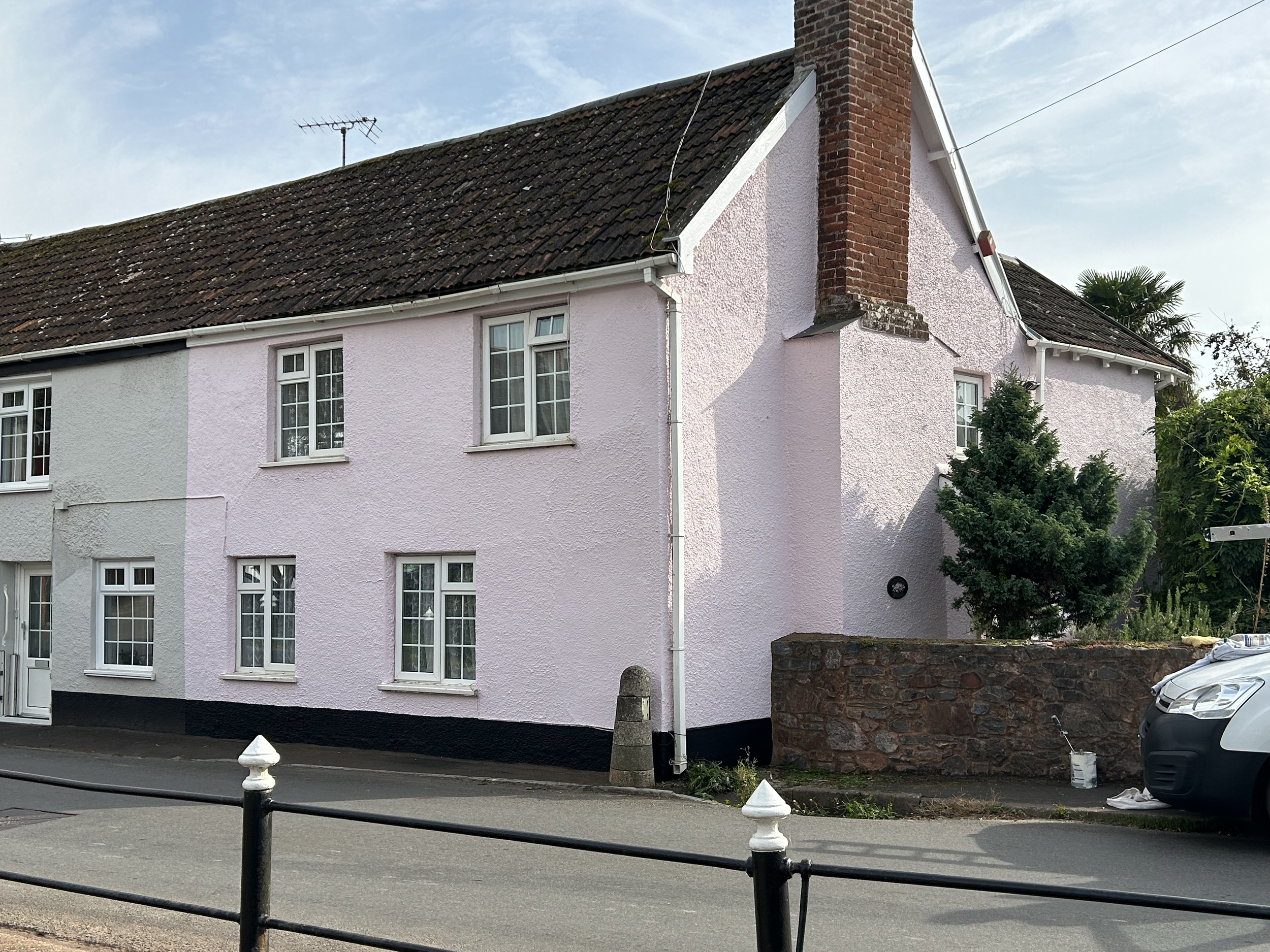 Pink two-story house with white window frames and a brick chimney, located on a street with a black metal fence and parked white vehicle.