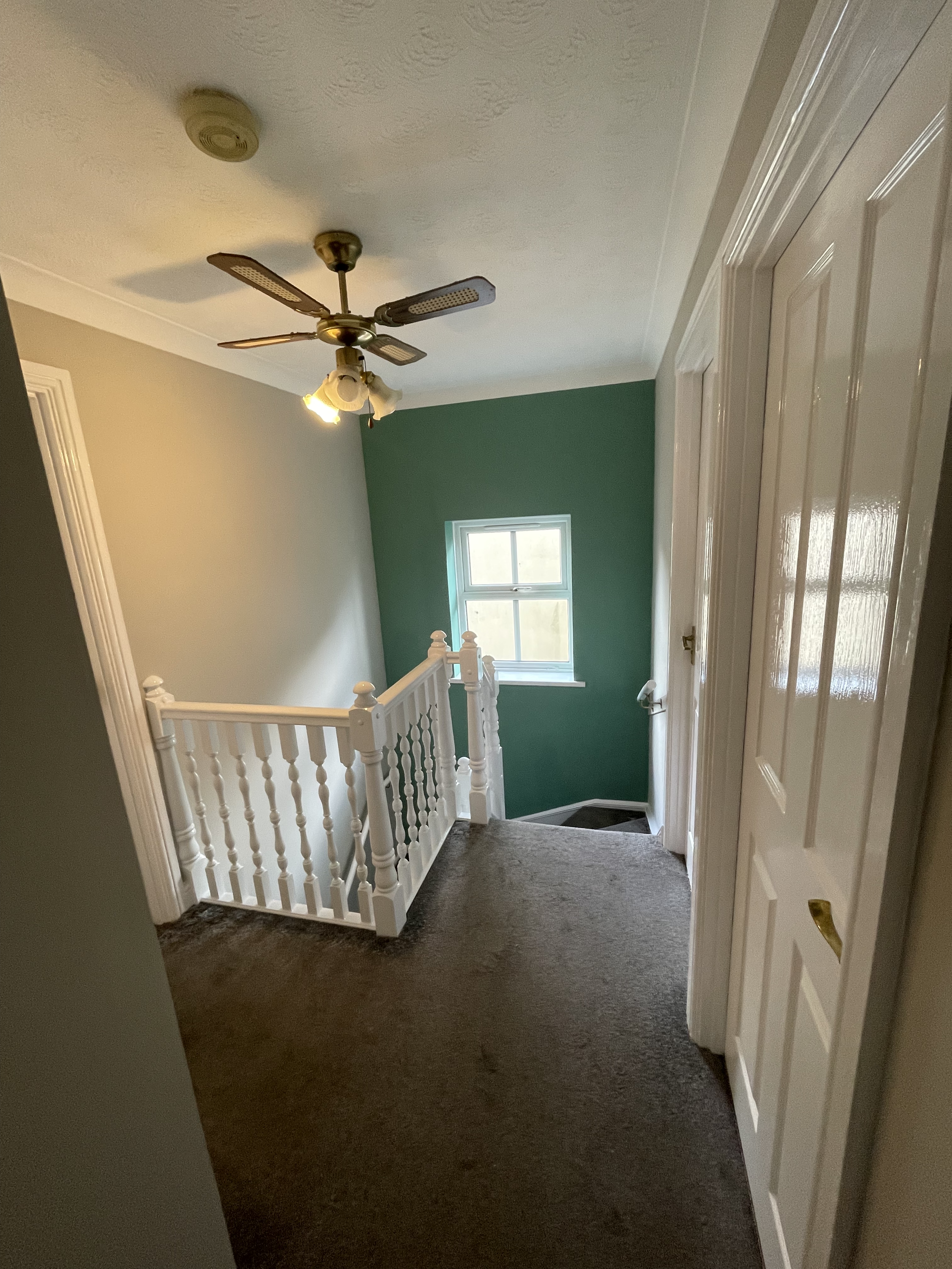 A house landing with a beige carpet, white railing, green accent wall, window, ceiling fan, and a white door.