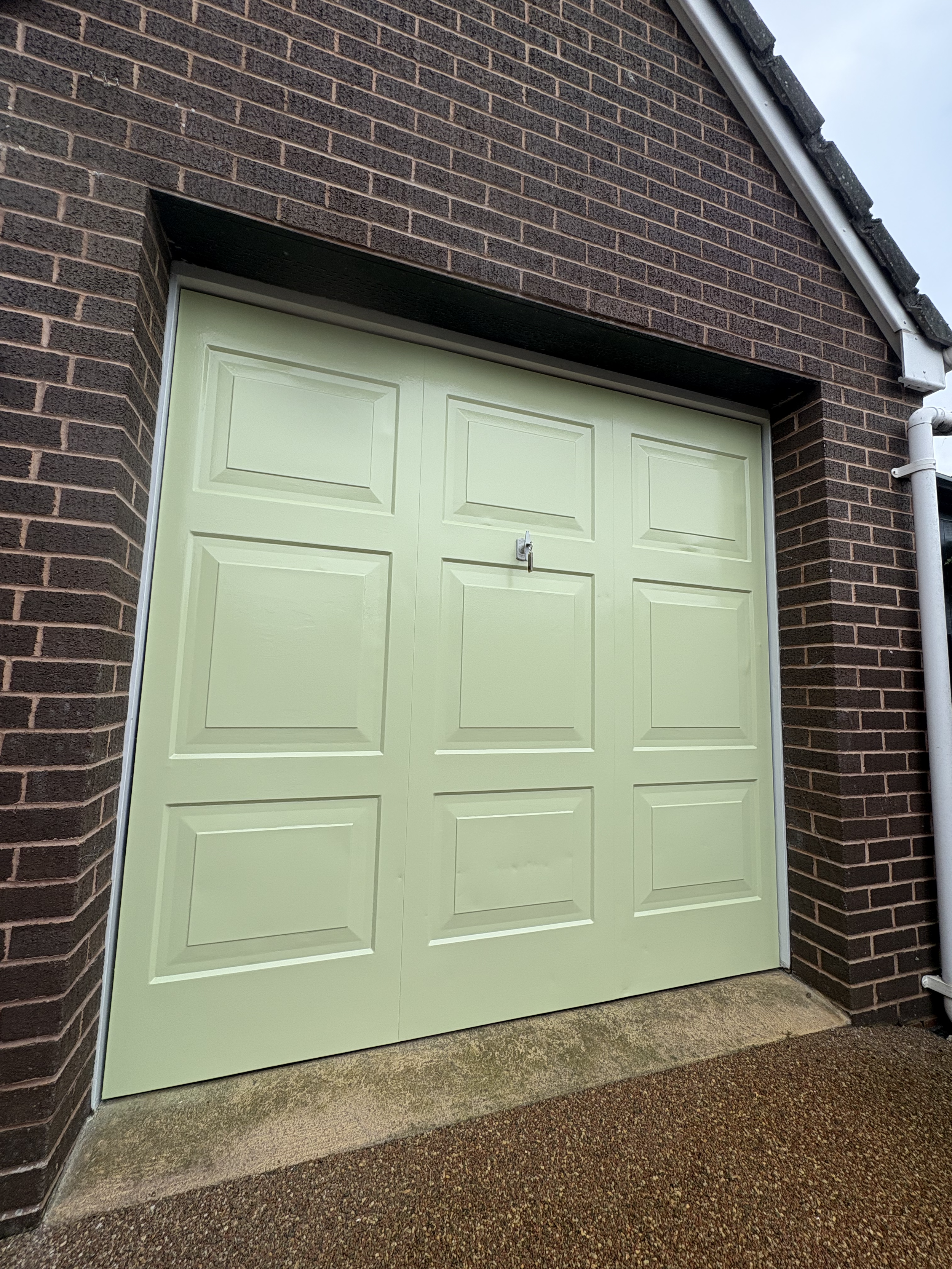 Light green garage door with a small padlock at the center, set in a brick wall with a sidewalk below.