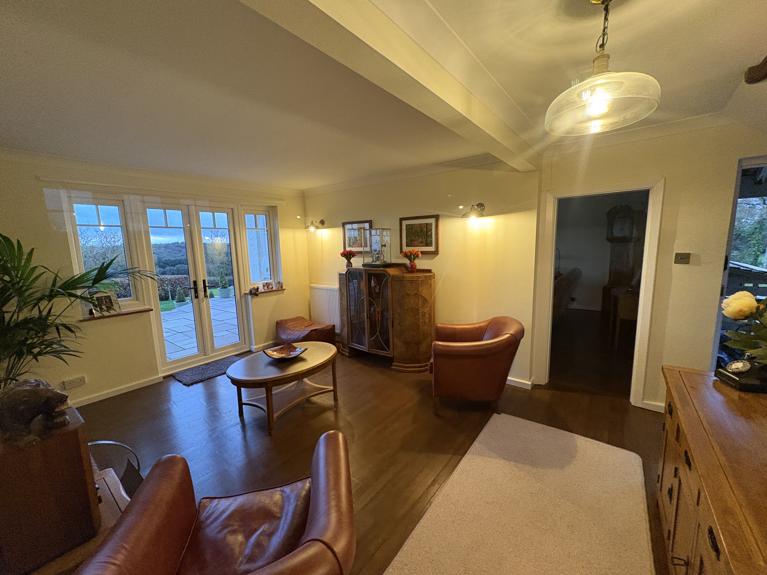 Living room with wooden flooring, leather armchairs, a glass coffee table, large windows with a sliding door opening to a patio, and a countryside view outside.