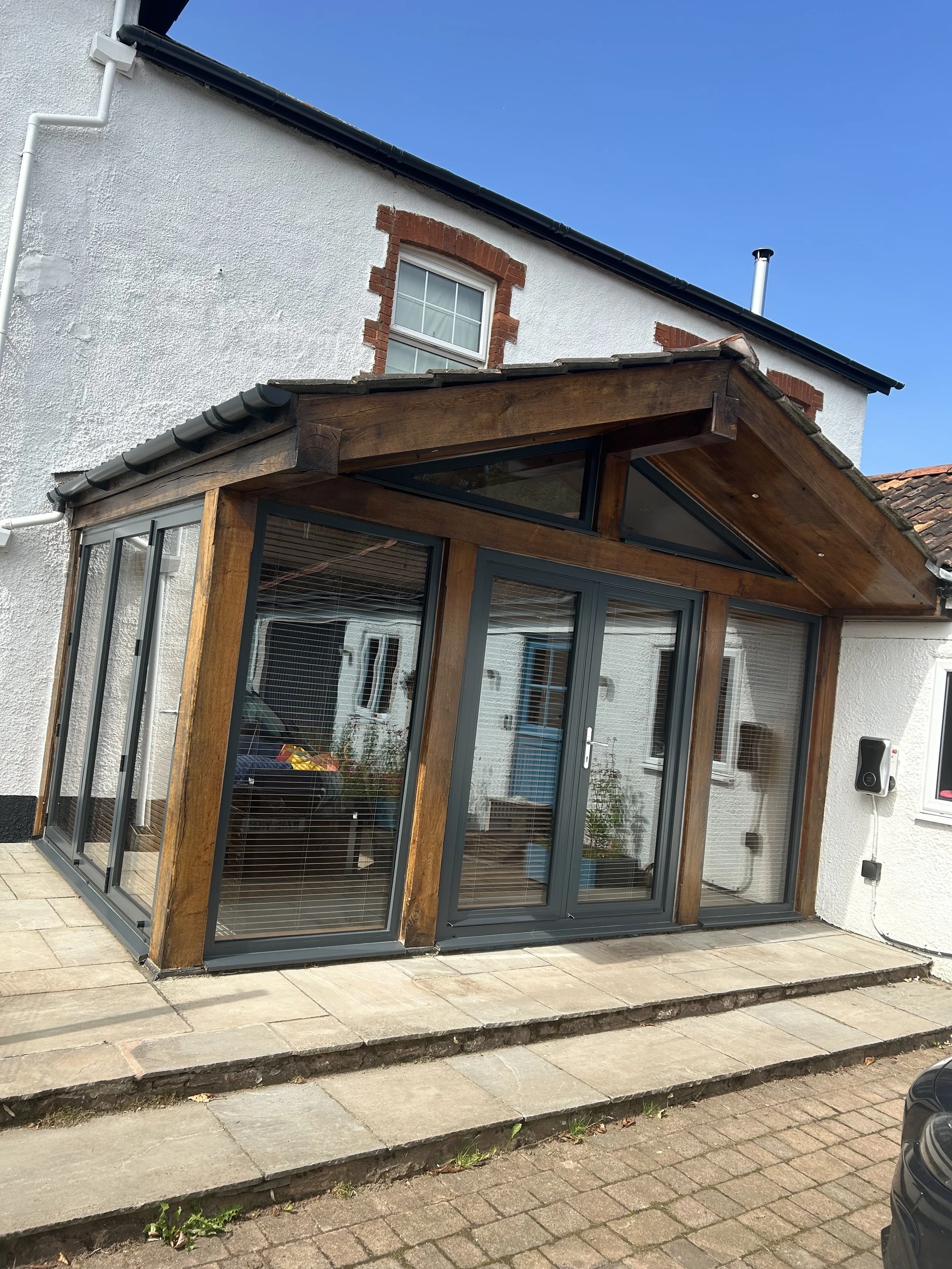 A sunroom extension with wooden framing and glass sliding doors and windows attached to a white house, with steps leading up to the entrance.