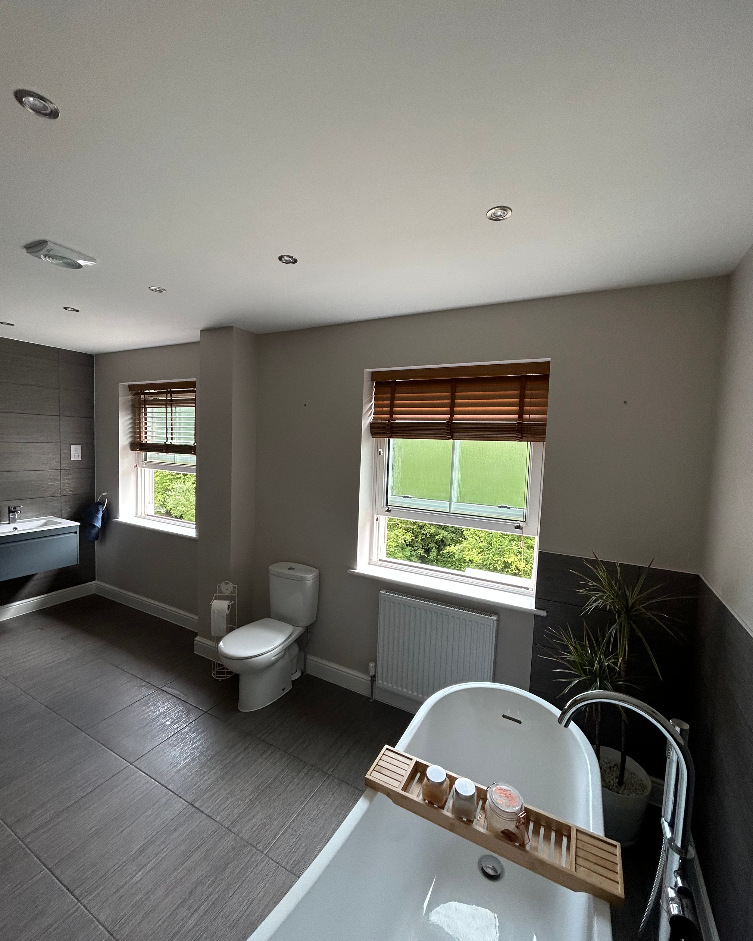 Modern bathroom with a bathtub near the window, a toilet, a sink with a mirror, and a potted plant. Wooden blinds cover the windows, and the floor is tiled.