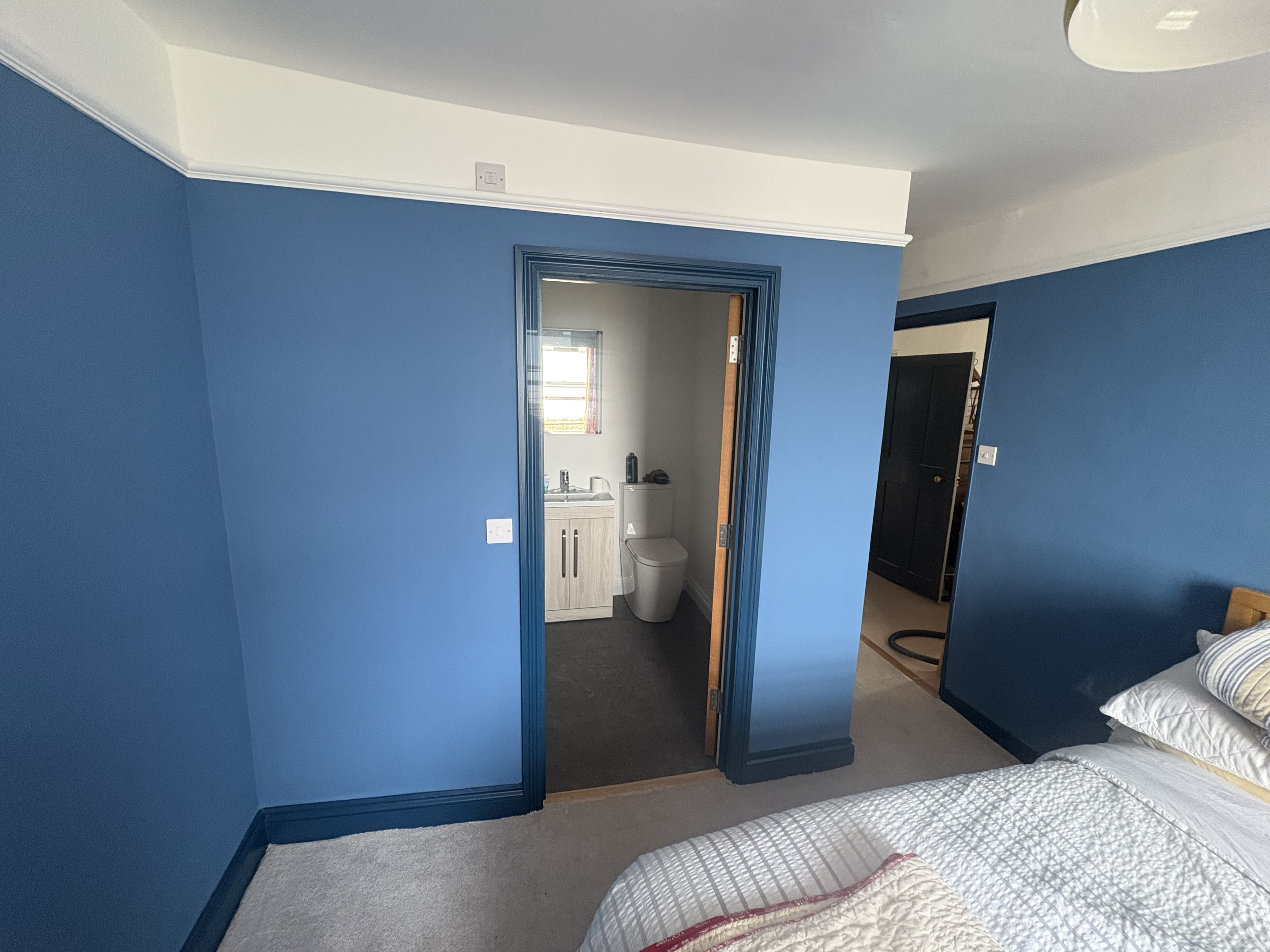A bedroom with blue walls and trim, partially visible bed with patterned bedding, and a doorway leading to a small bathroom. The bathroom has a window with blinds, a sink with a cabinet, and a toilet.