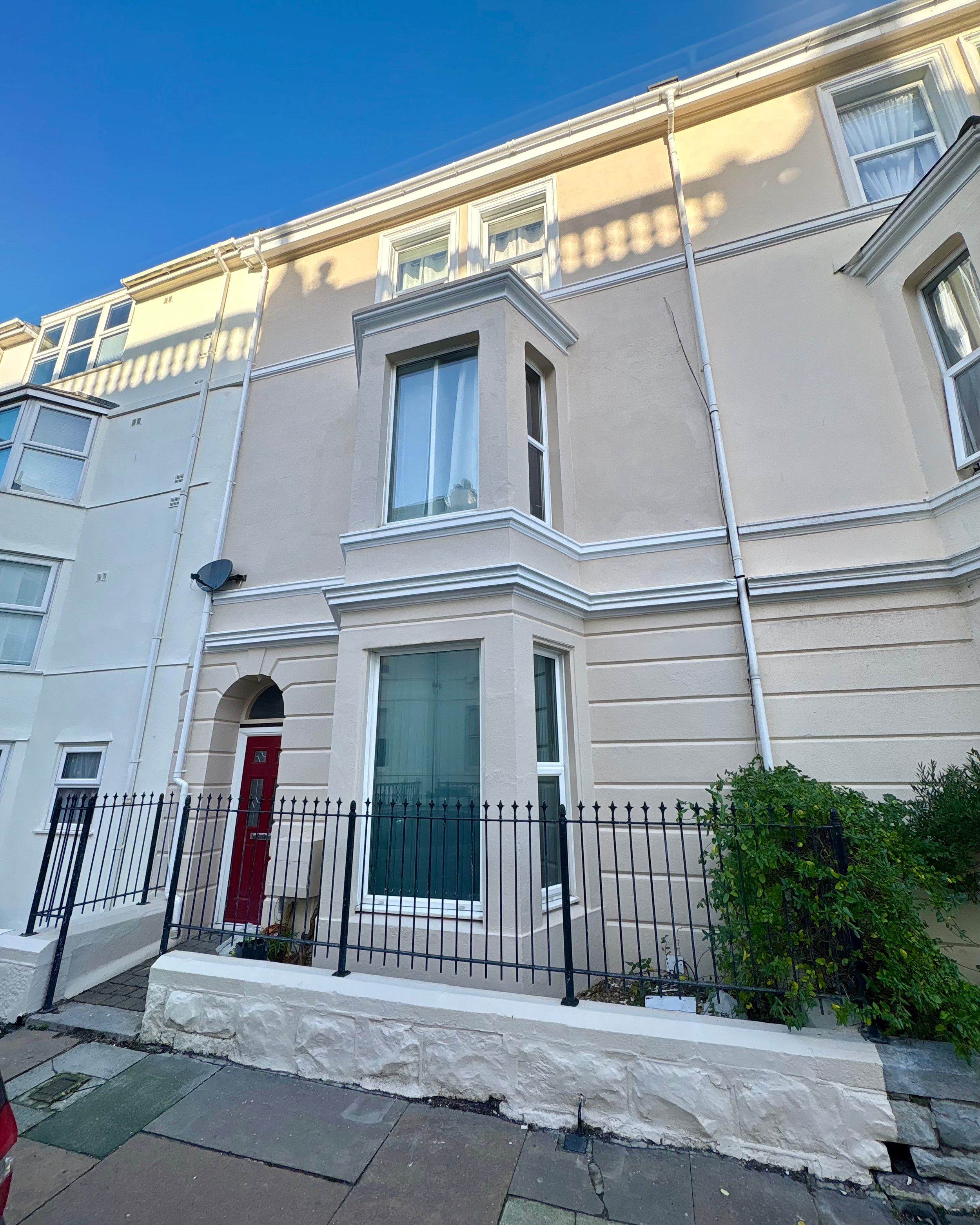 Front view of a white three-story residential building with bay windows, a red front door, a small fenced porch, and a satellite dish on the wall, under a clear blue sky.
