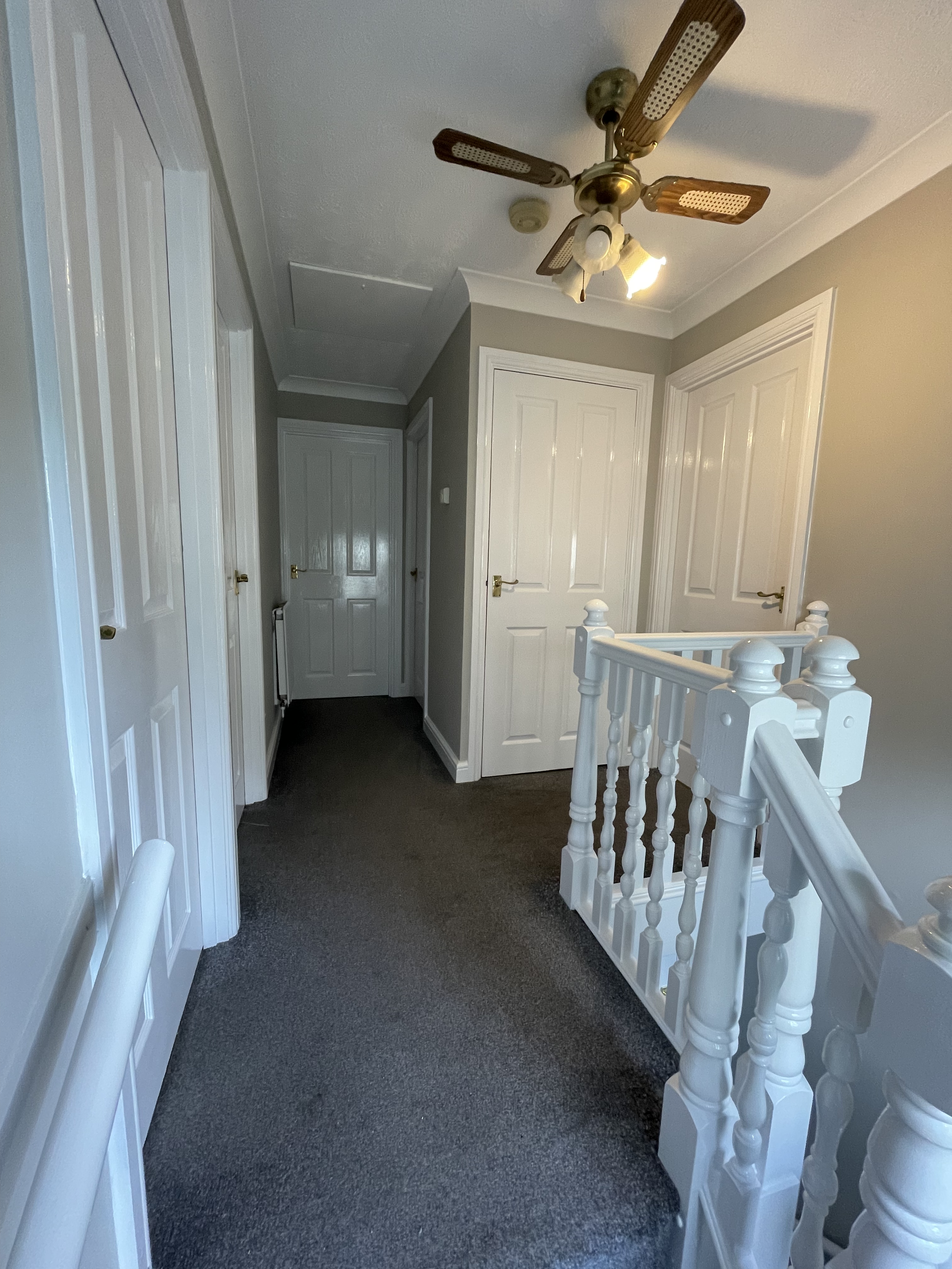 A hallway with white paneled doors, a white wooden staircase railing, gray walls, a dark carpeted floor, and a ceiling fan with wooden blades and a light fixture.