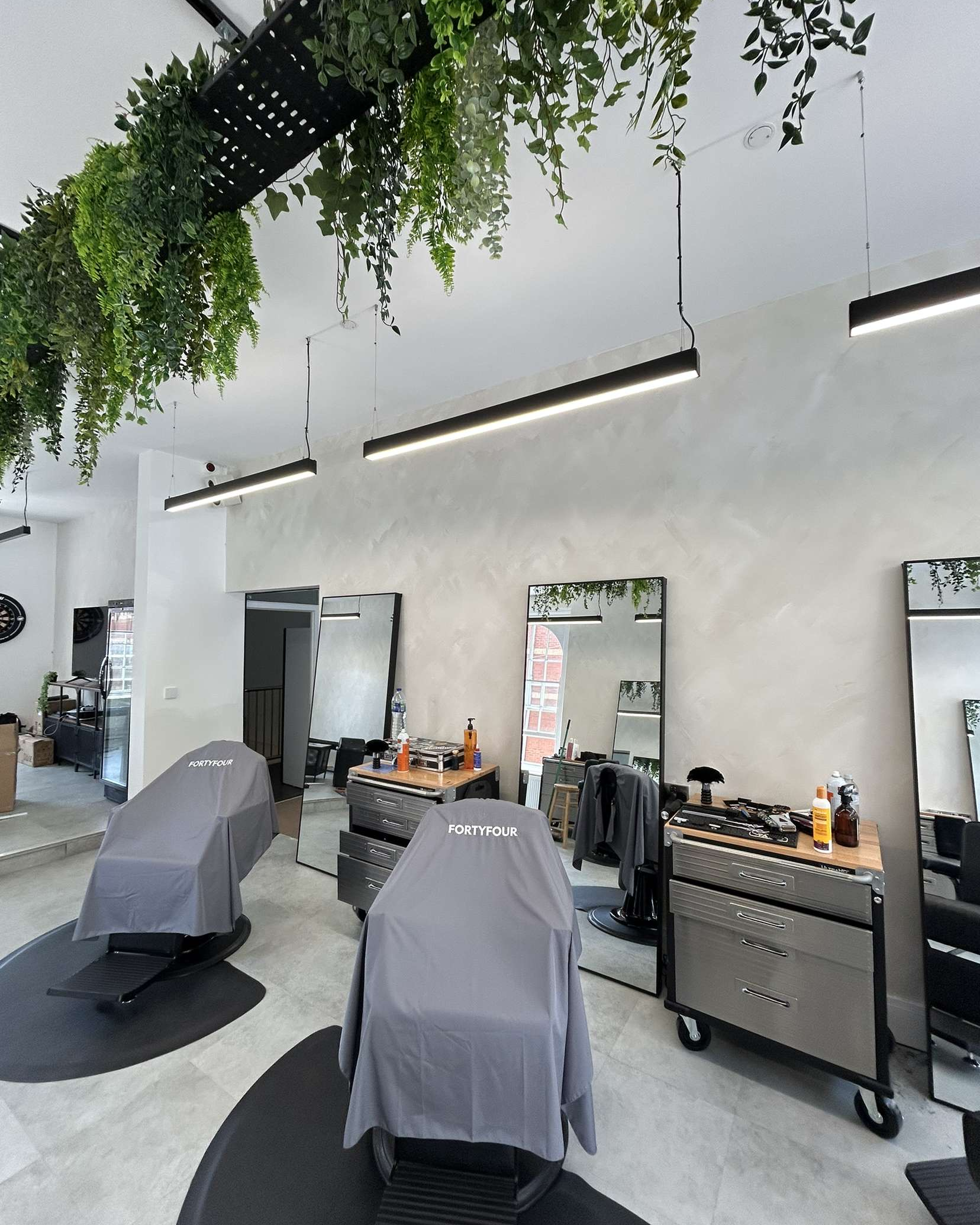 Inside a modern barbershop with two barber chairs covered with gray cloths, mirrors, tools on carts, and hanging ceiling lights, with greenery hanging from the ceiling.