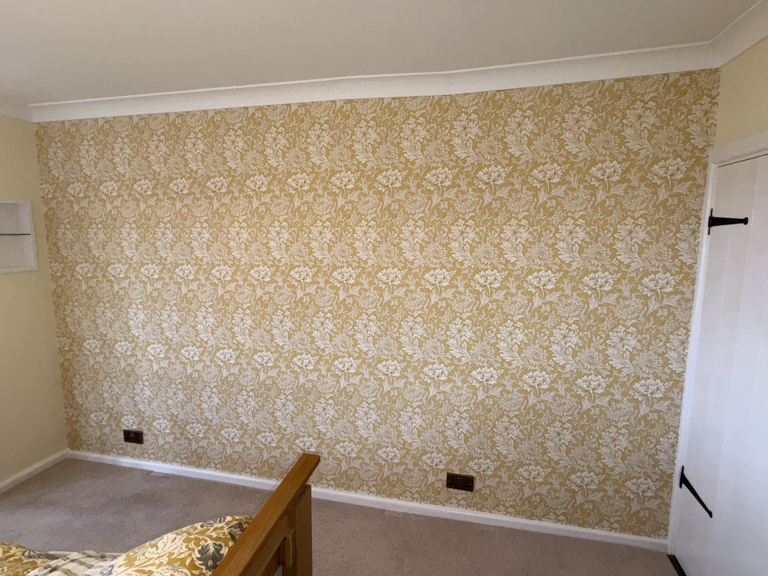 A decorated bedroom wall with yellow floral damask wallpaper, beige carpet, a wooden bed frame with a floral bedspread, white crown molding, electrical outlets, and a white door with black hinges.