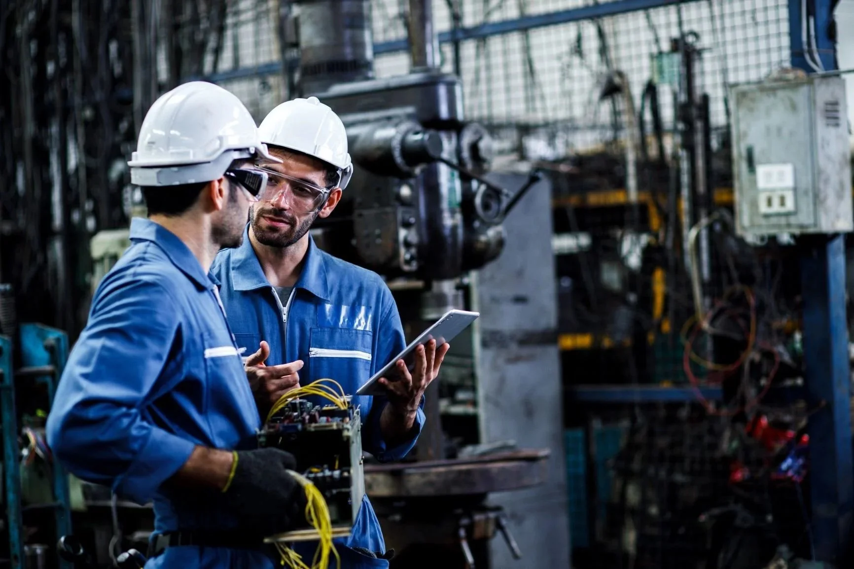 Two male workers in safety helmets and blue uniforms discussing in an industrial facility with machinery in the background.