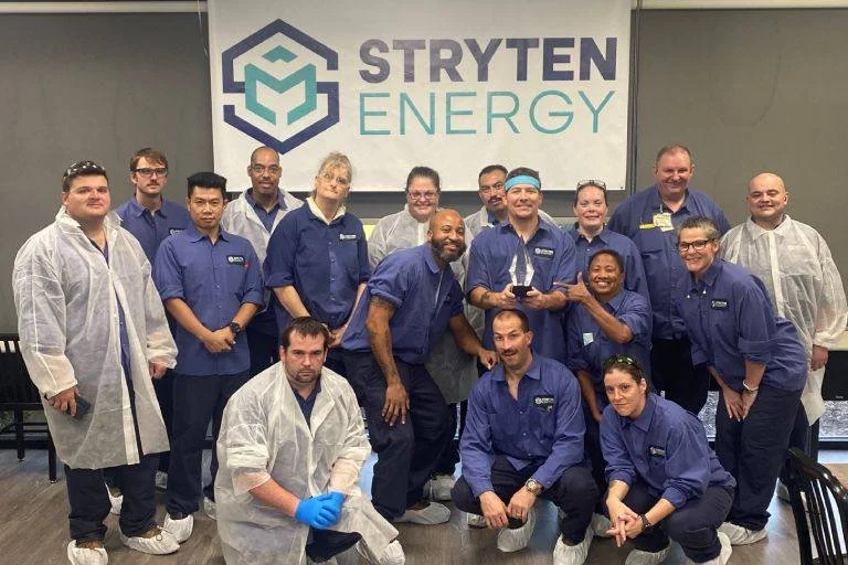 Group of employees from Stryten Energy posing indoors, some wearing lab coats and gloves, in front of a Stryten Energy banner with logo, smiling and gathered for a team photo.
