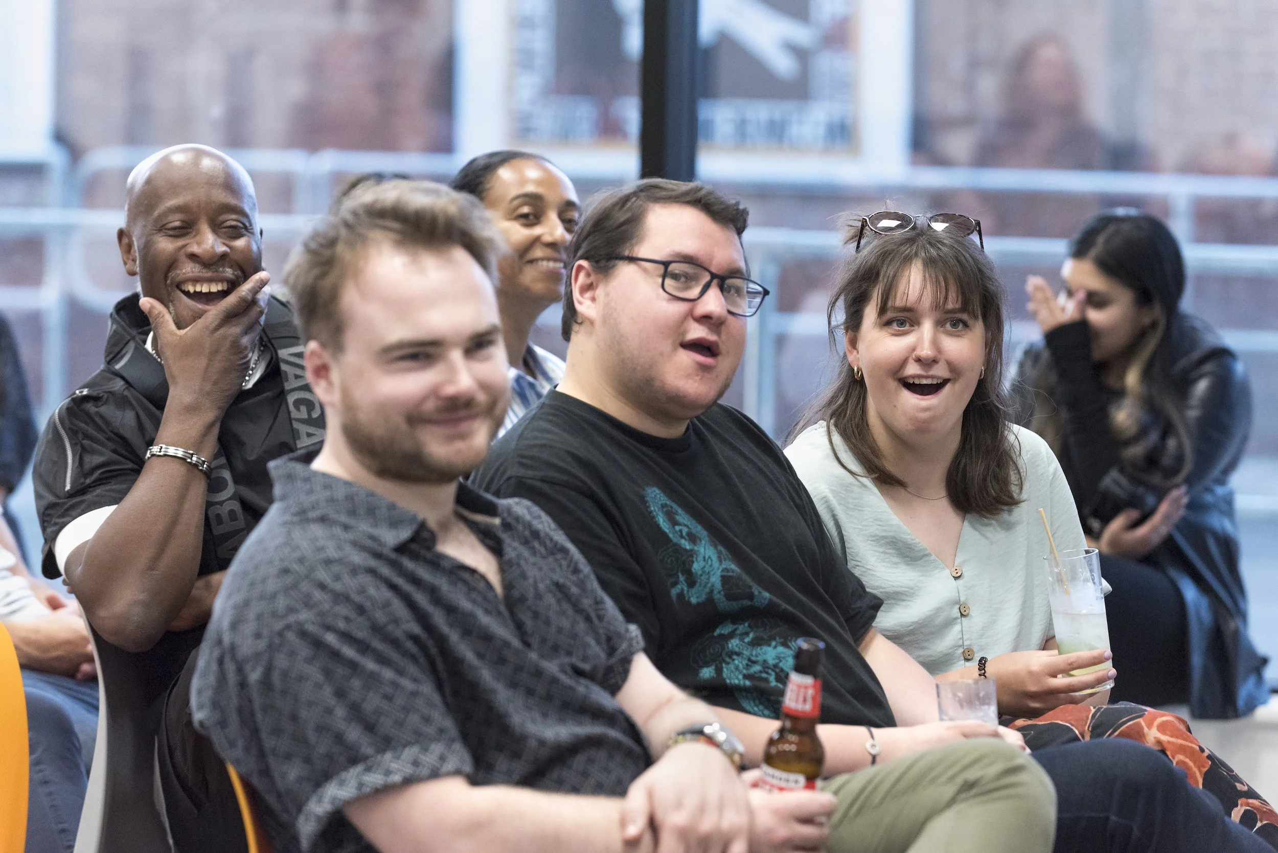 Group of diverse people sitting indoors, enjoying a social event, some laughing and smiling, with drinks in hand.