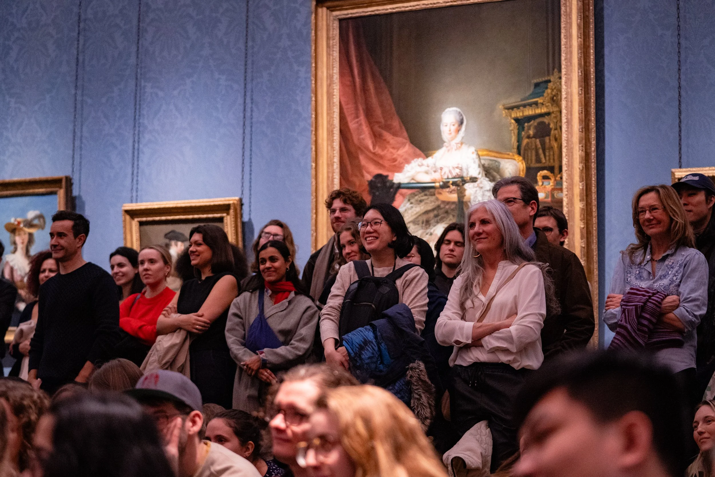 A group of people standing and smiling inside an art museum, with large framed paintings hanging on blue patterned walls behind them.
