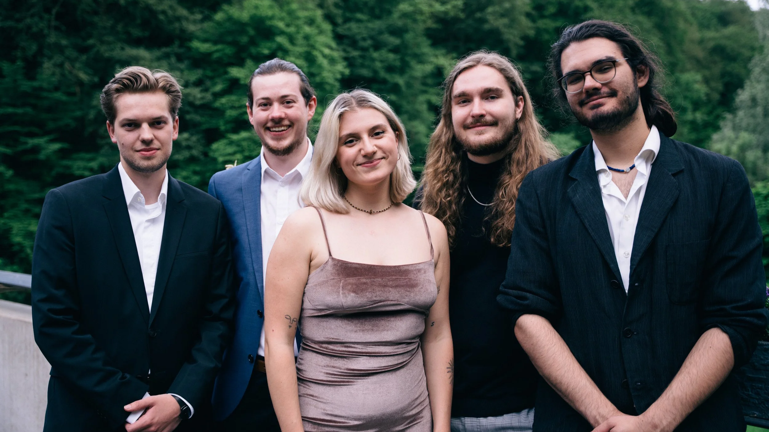 Fünf junge Menschen stehen im Freien vor einer grünen Waldkulisse, lächeln und posieren für ein Gruppenfoto.