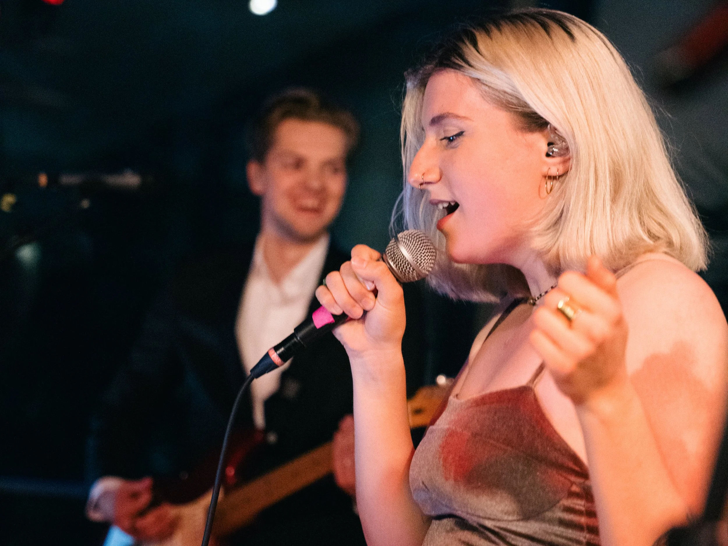 Eine blonde Sängerin mit einem Mikrofon singt auf einer Bühne, während ein Mann im Hintergrund Gitarre spielt.
