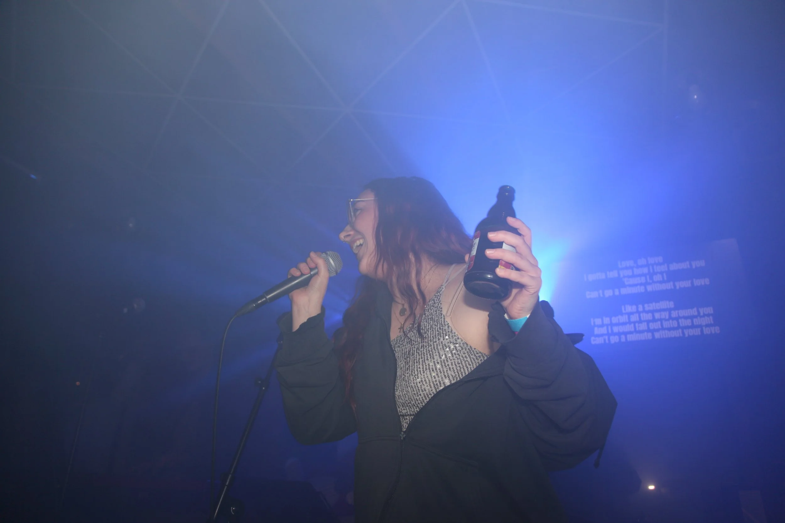Eine weibliche Person singt in einem dunklen Raum mit blauem Licht, hält eine Mikrofon und ein Bierflasche, während sie lächelt.
