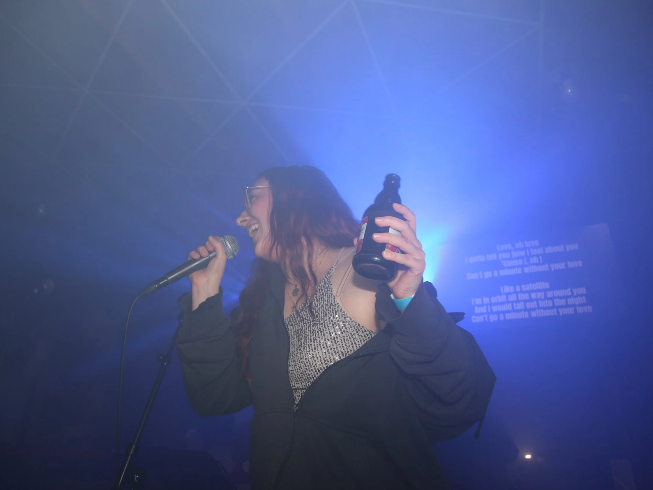 Eine Frau mit Brille singt in einem dunklen Raum mit bläulichem Licht, hält eine Flasche Bier in der Hand, während sie in ein Mikrofon singt.