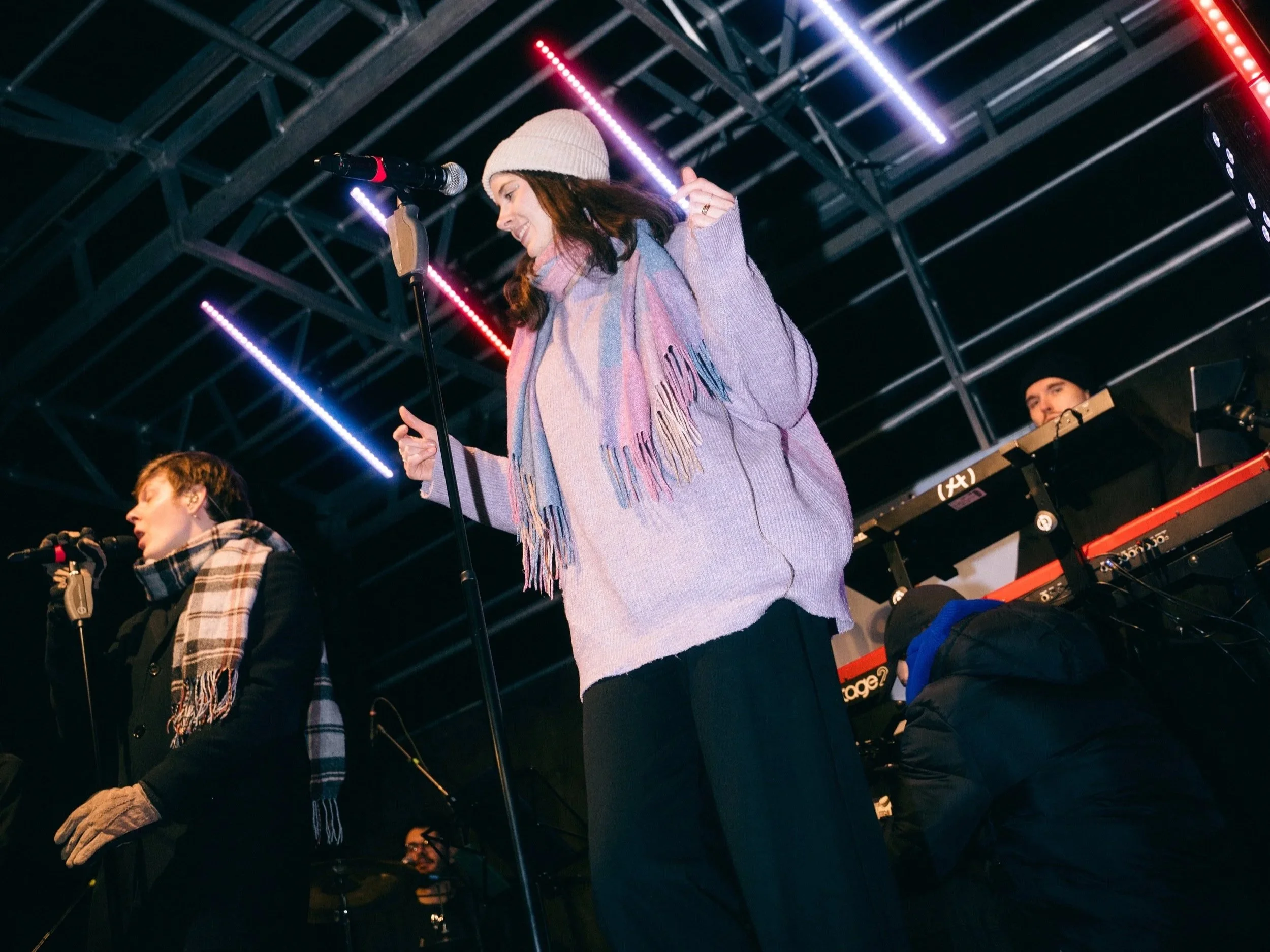 Drei Musiker performen auf einer Bühne bei Nacht, eine Frau mit Mütze und Schal, hinter einem Keyboard spielt ein Mann, eine weitere Person steht im Vordergrund, alle in Winterkleidung.