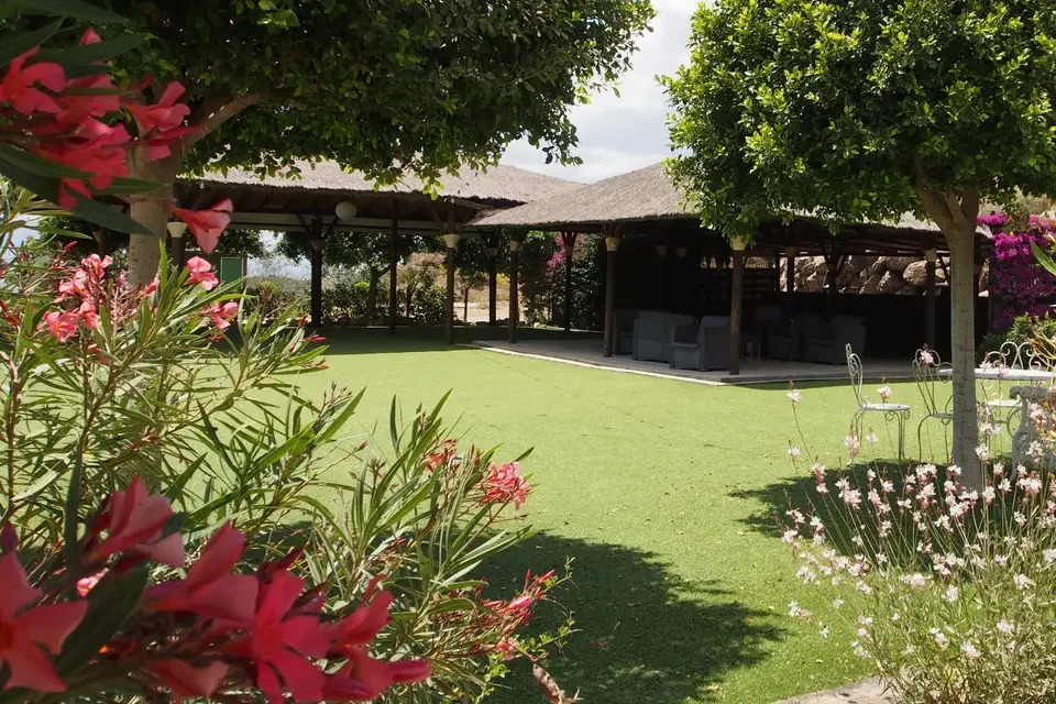 Jardín con césped, plantas con flores rosadas y árboles verdes, área de descanso con sombra y muebles en un espacio exterior.